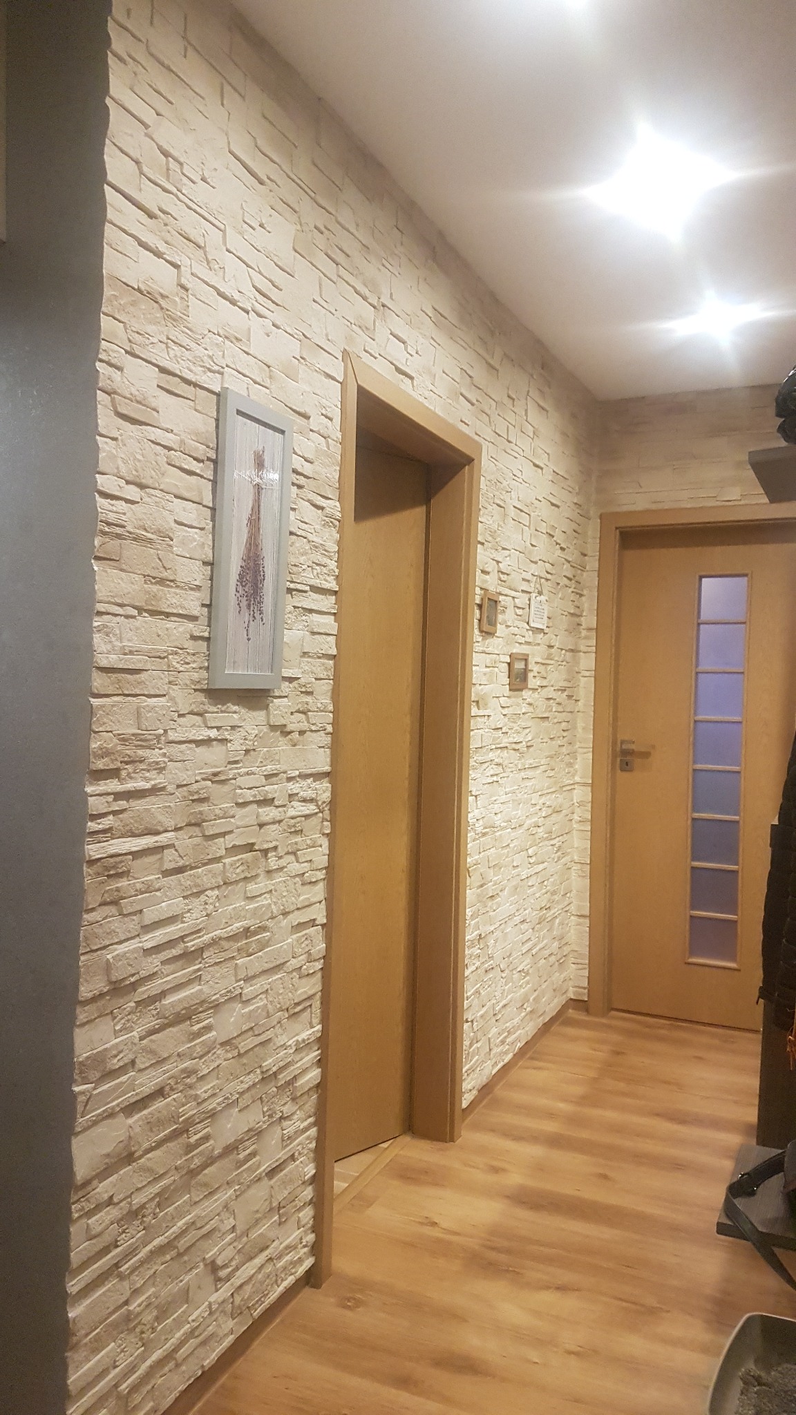 Korytarz z dekoracyjną ścianą z imitacji kamienia, drewnianą podłogą i dwiema drewnianymi ościeżnicami, na ścianie wisi obrazek w szarej ramce.