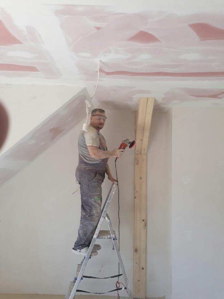 Pracownik na drabinie szlifuje sufit pokryty gładzią, używając szlifierki elektrycznej; widoczne ślady po łatach na suficie, okulary ochronne na twarzy pracownika.