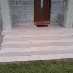 Wyłożone jasnymi płytkami schody wejściowe do domu z brązowymi drzwiami, surowy stan ścian z bloczków betonowych i częściowo otynkowanymi kolumnami.