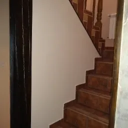 Wykończone schody wewnętrzne z brązowymi płytkami imitującymi kamień, biała ściana, drewniana balustrada z dekoracyjnymi elementami, widok z dołu.