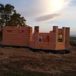 Wznoszenie ścian parterowego budynku z czerwonej ceramiki poryzowanej, widoczny pracownik w odblaskowej kamizelce, częściowo ułożone bloczki, teren budowy w tle.
