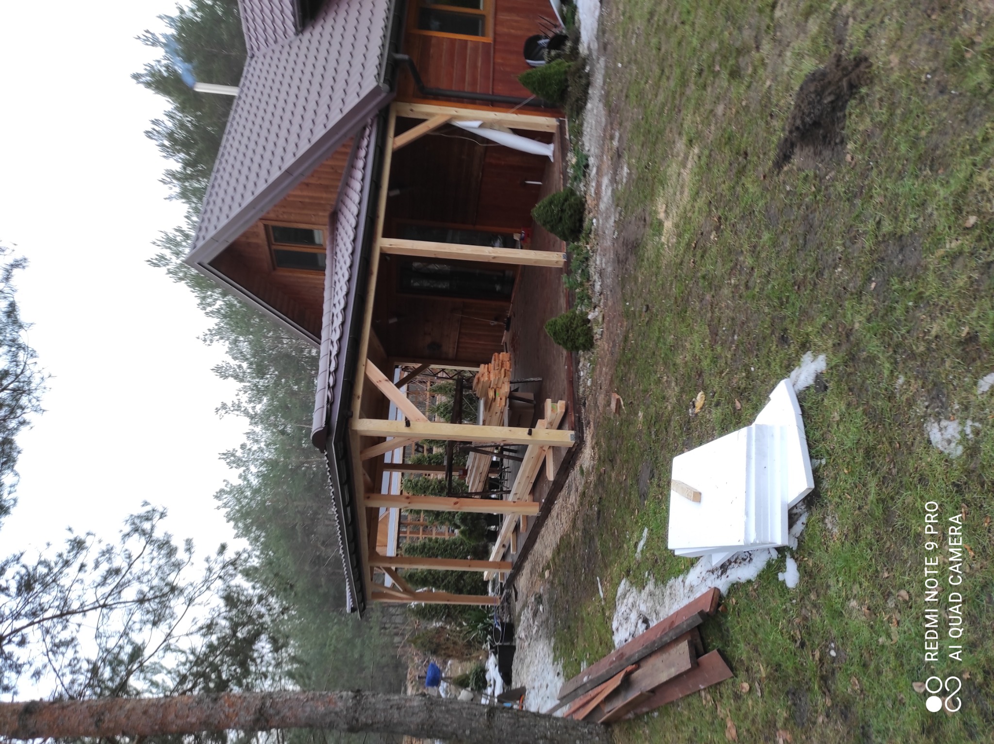 Dom z drewnianą werandą w trakcie budowy, widoczne elementy konstrukcyjne, deski i materiały budowlane na trawie.