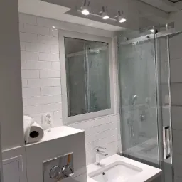 Nowoczesna, jasna łazienka z białymi, prostokątnymi płytkami, kabiną prysznicową ze szklanymi drzwiami i umywalką wpuszczoną w blat z baterią. Widoczny fragment toalety z chromowanym przyciskiem...