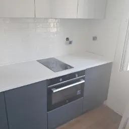 Fragment nowej kuchni z szarymi szafkami dolnymi, białymi górnymi, białym blatem, płytą indukcyjną i piekarnikiem Electrolux; na ścianie białe płytki typu metro, widoczne puszki elektryczne.