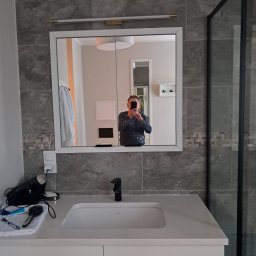 Janusz Kowalski - Wykończona łazienka z umywalką nablatową, szarą płytką imitującą kamień i kabiną prysznicową typu walk-in. W lustrze odbija się osoba robiąca zdjęcie smartfonem, ubrana w robocze ubranie z plamami...