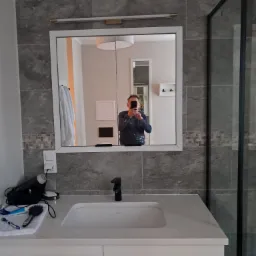 Wykończona łazienka z umywalką nablatową, szarą płytką imitującą kamień i kabiną prysznicową typu walk-in. W lustrze odbija się osoba robiąca zdjęcie smartfonem, ubrana w robocze ubranie z plamami...