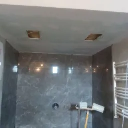 Łazienka w trakcie prac: ściany wyłożone szarymi płytkami imitującymi marmur, sufit po szpachlowaniu z widocznymi otworami na oświetlenie, grzejnik drabinkowy i narzędzia glazurnicze.