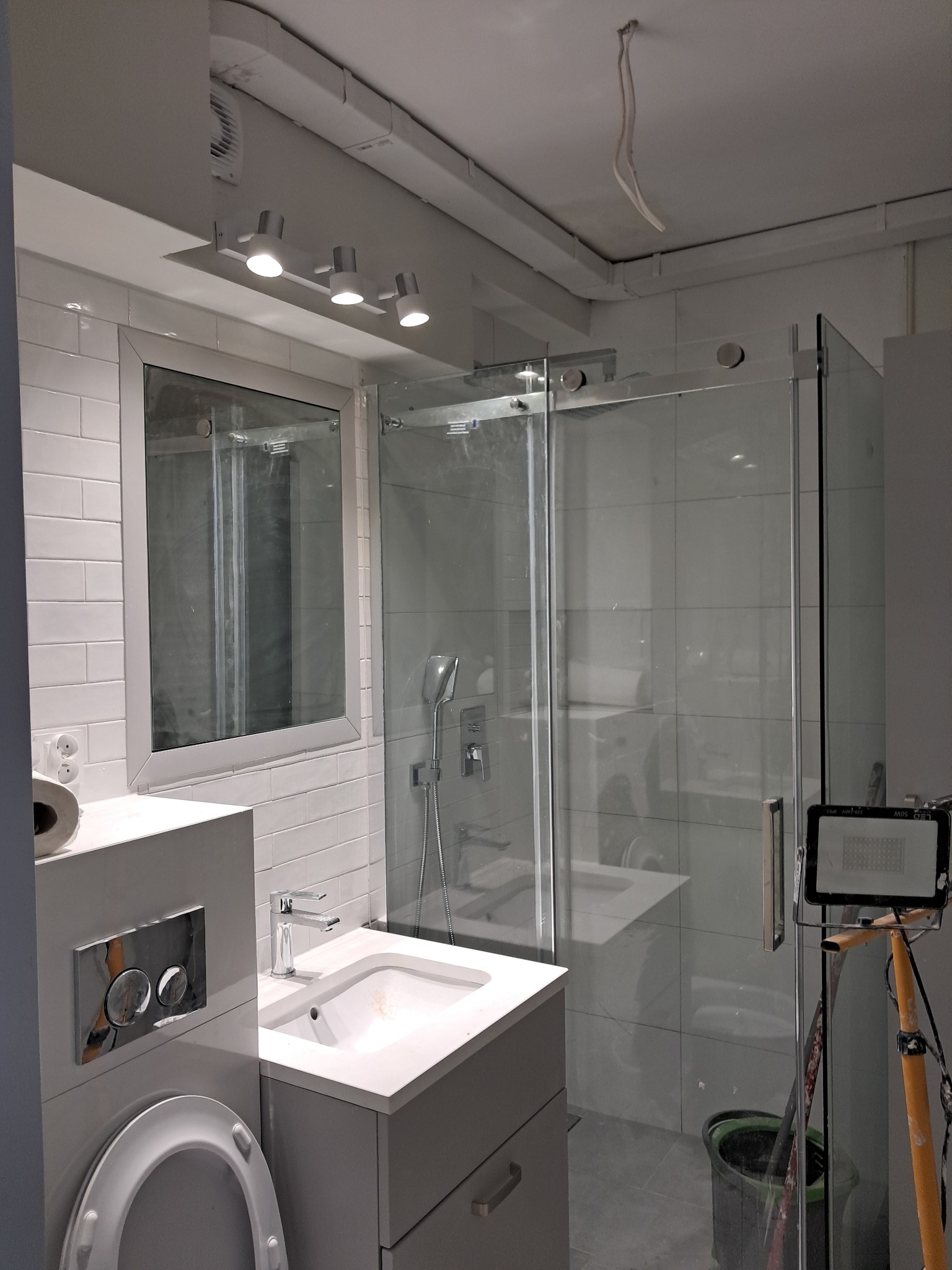 Nowoczesna łazienka w trakcie wykańczania: białe płytki imitujące cegłę, szklana kabina prysznicowa, umywalka nablatowa z chromowaną baterią, podwieszana toaleta z przyciskiem spłukującym. Widoczna...