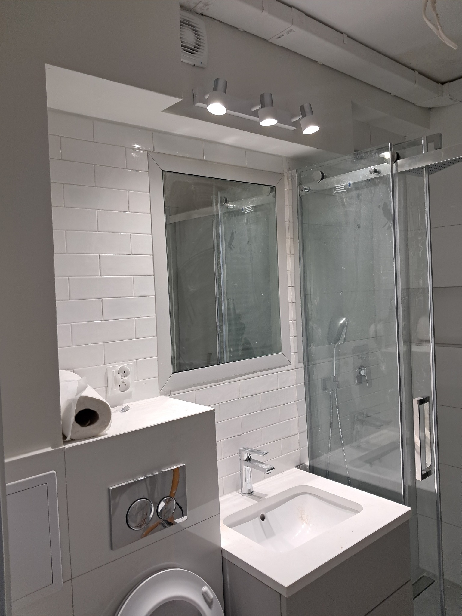 Nowoczesna, jasna łazienka z białymi, prostokątnymi płytkami, kabiną prysznicową ze szklanymi drzwiami i umywalką wpuszczoną w blat z baterią. Widoczny fragment toalety z chromowanym przyciskiem...