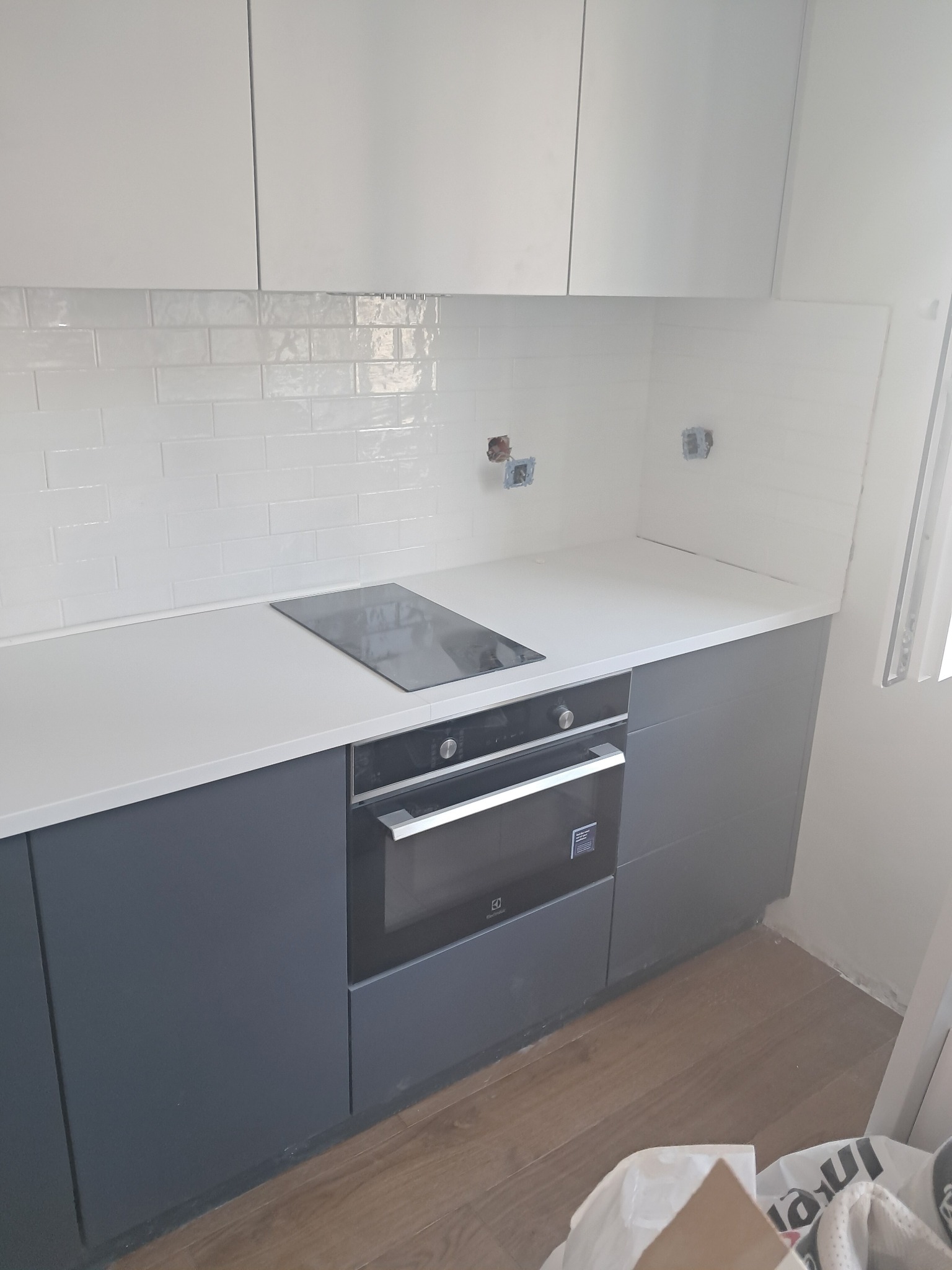 Fragment nowej kuchni z szarymi szafkami dolnymi, białymi górnymi, białym blatem, płytą indukcyjną i piekarnikiem Electrolux; na ścianie białe płytki typu metro, widoczne puszki elektryczne.