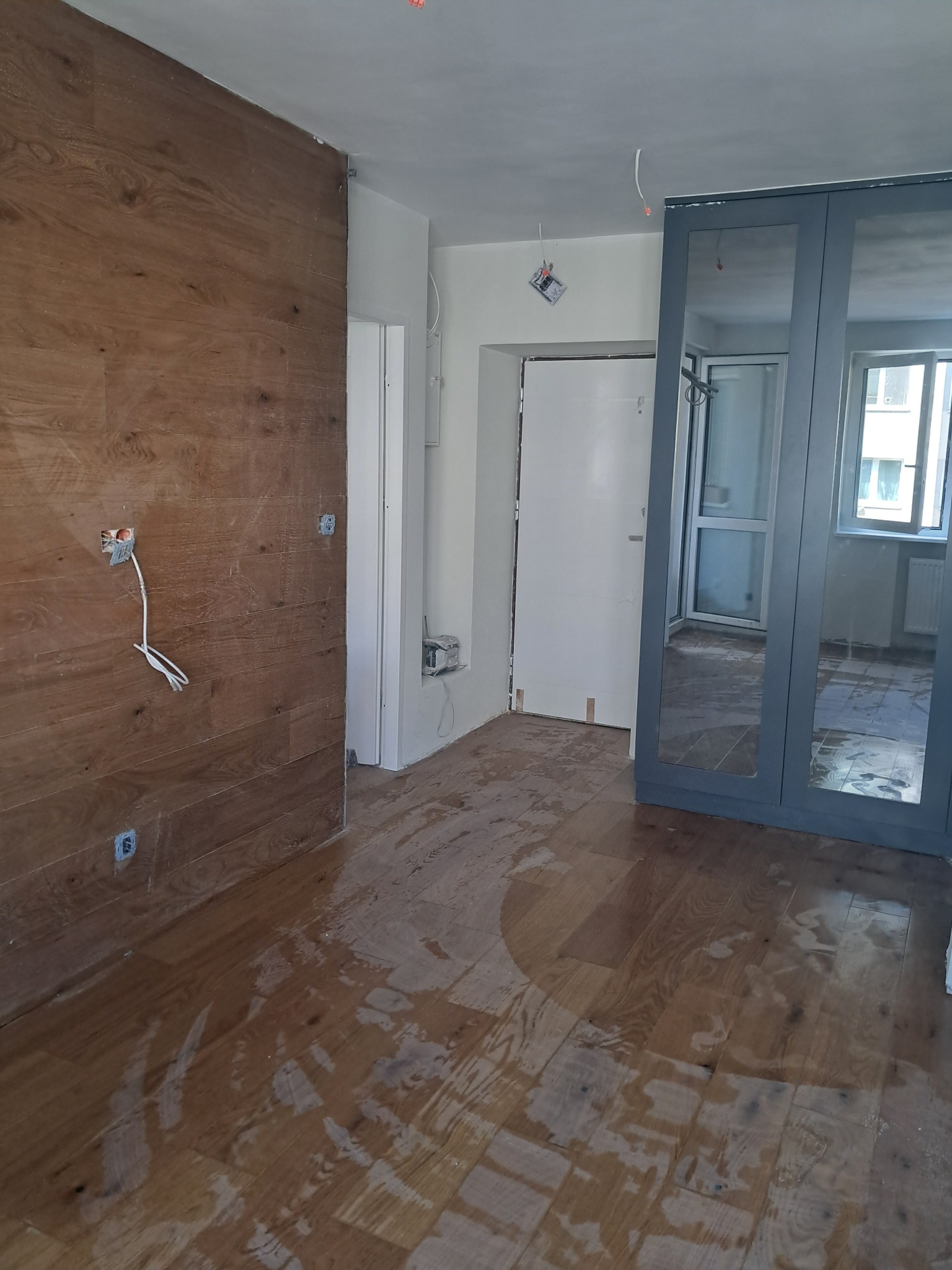 Wnętrze w trakcie prac wykończeniowych: drewniana okładzina ścienna, panele podłogowe z widocznymi śladami wilgoci, częściowo zamontowana szafa z lustrami i wystające kable elektryczne ze ściany.
