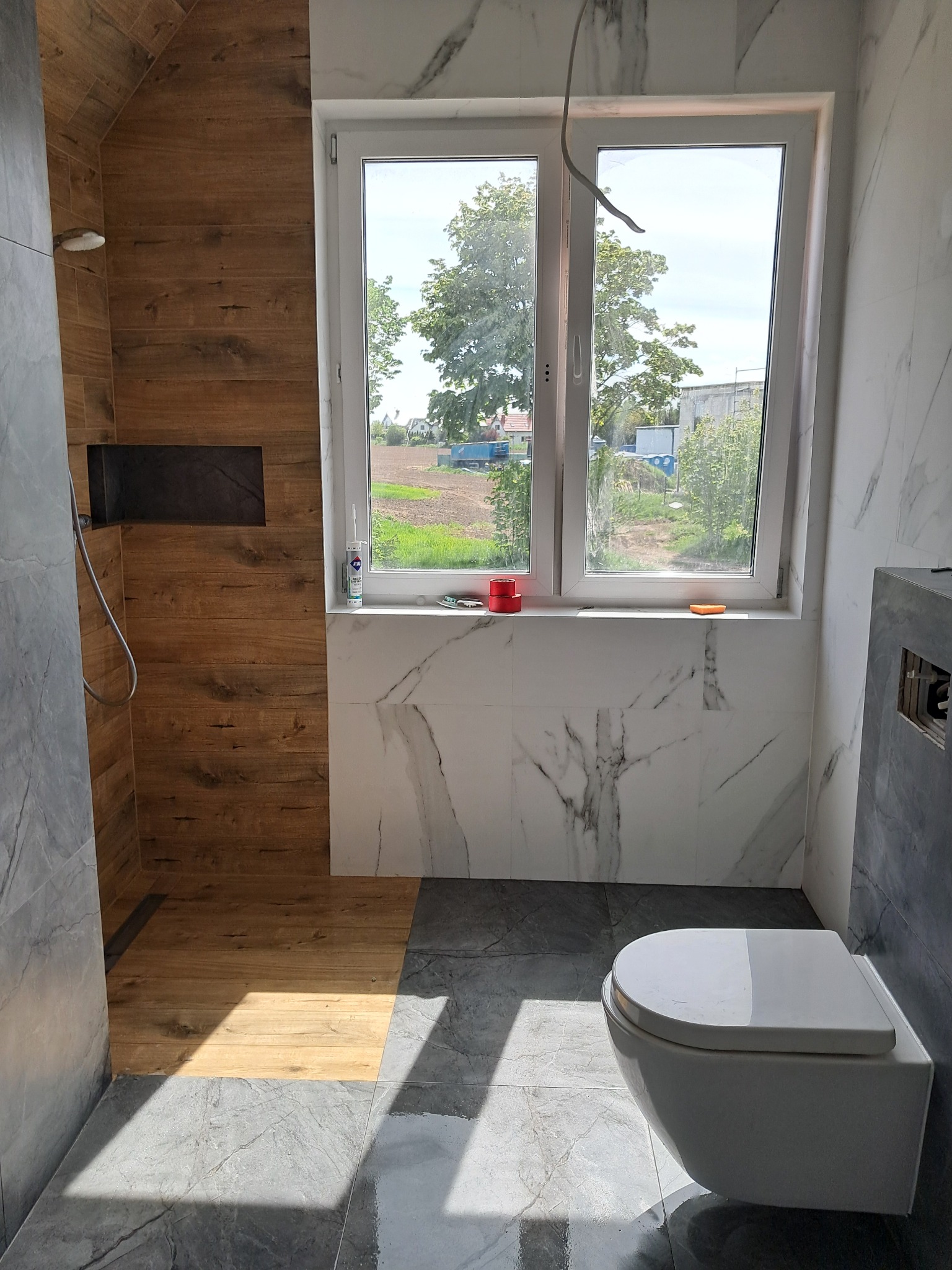 Nowoczesna łazienka z drewnopodobnymi płytkami w strefie prysznicowej, marmurowymi płytkami na ścianach i szarymi płytkami na podłodze, z podwieszaną toaletą i oknem z widokiem na zewnątrz.