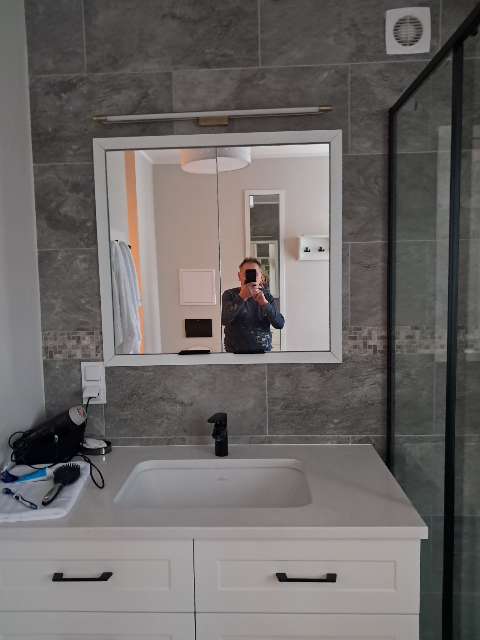 Wykończona łazienka z umywalką nablatową, szarą płytką imitującą kamień i kabiną prysznicową typu walk-in. W lustrze odbija się osoba robiąca zdjęcie smartfonem, ubrana w robocze ubranie z plamami...