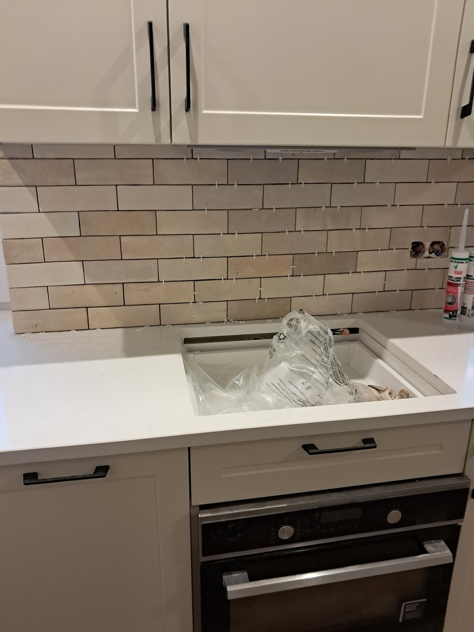 Ściana nad blatem kuchennym z ułożonymi beżowymi płytkami w trakcie fugowania, widoczne krzyżyki dystansowe, biały blat i szafki z czarnymi uchwytami, puszki elektryczne bez osprzętu.