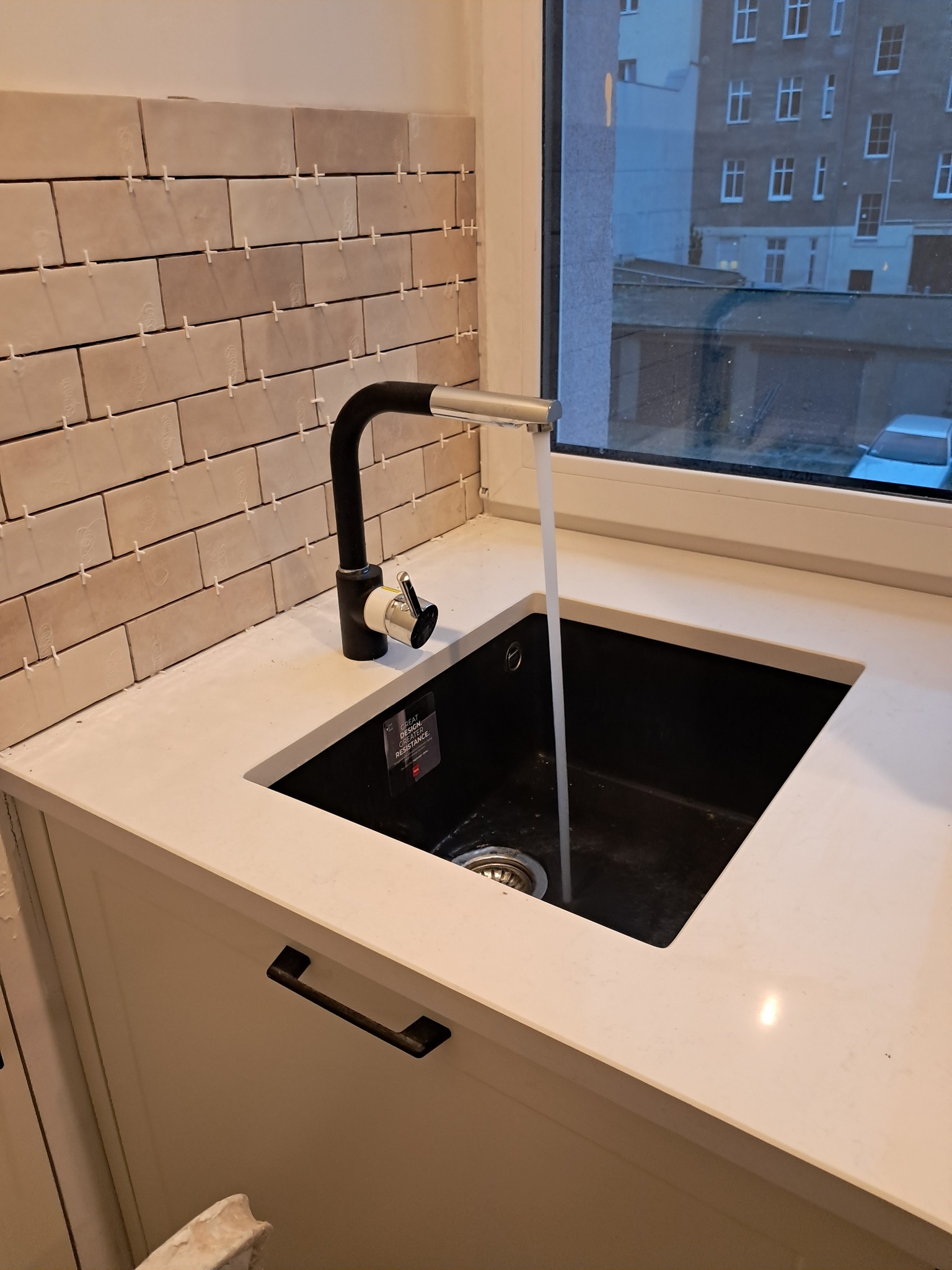 Montaż beżowych, prostokątnych płytek na ścianie nad czarnym zlewem z baterią, widoczne krzyżyki dystansowe, w tle okno z widokiem na budynek.