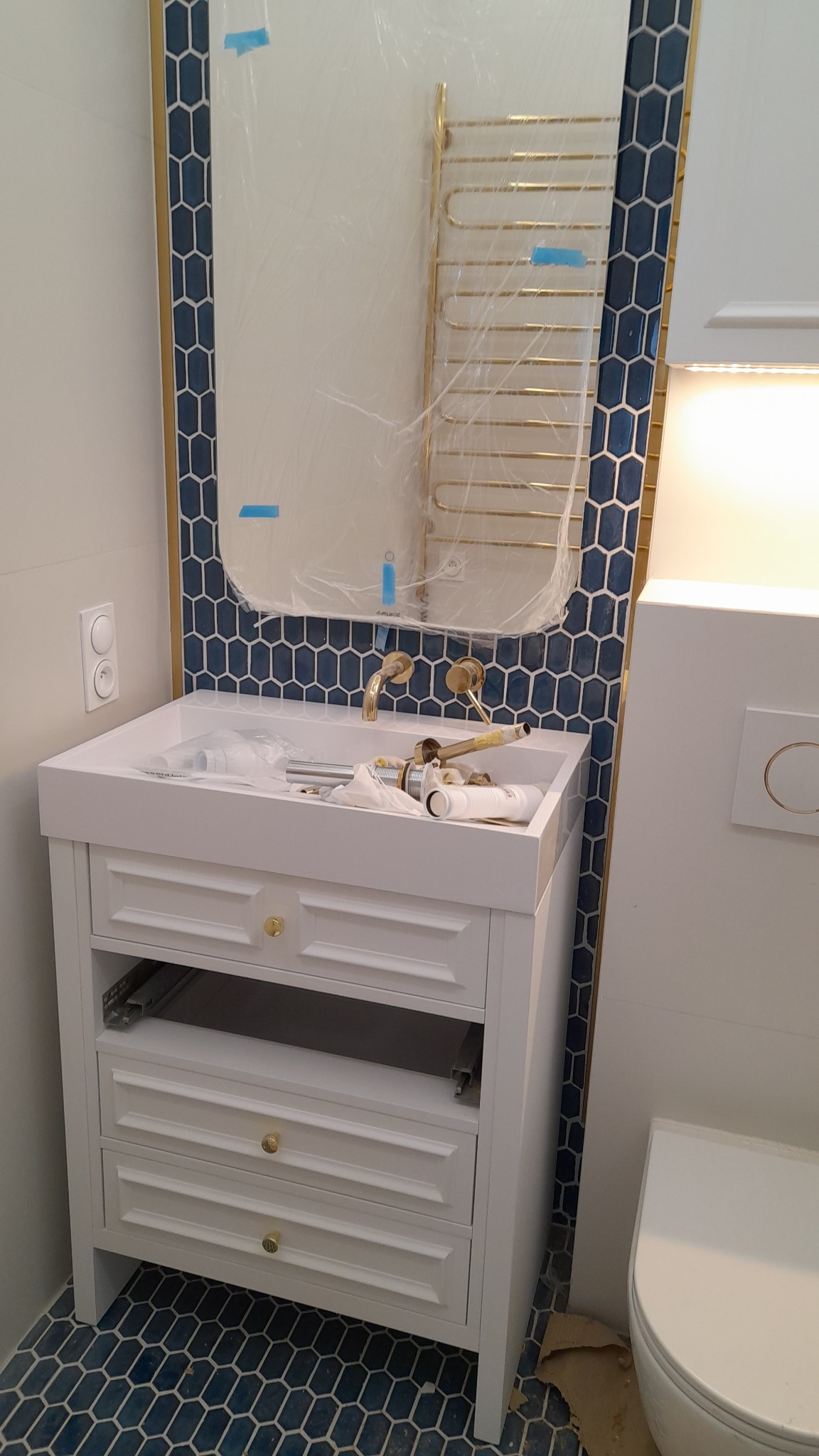 Montaż umywalki nablatowej z szafką w łazience z niebieską mozaiką heksagonalną na ścianie za lustrem w folii ochronnej i na podłodze, widoczne elementy instalacji wodnej i biała toaleta.