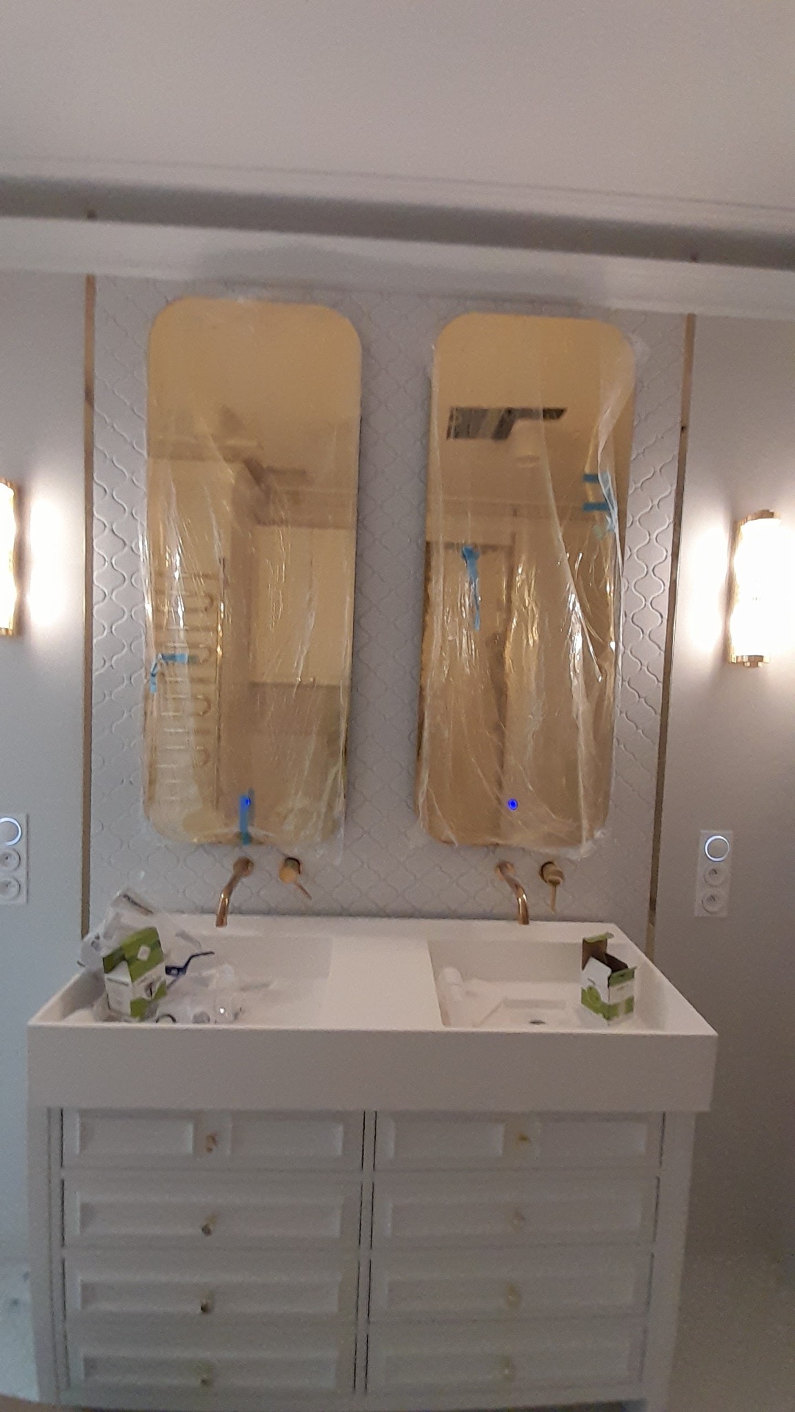 Biała szafka pod umywalkę z dwiema umywalkami i złotymi bateriami, nad którymi wiszą dwa podłużne lustra zabezpieczone folią ochronną, na ścianie wzorzyste płytki i dwa kinkiety.