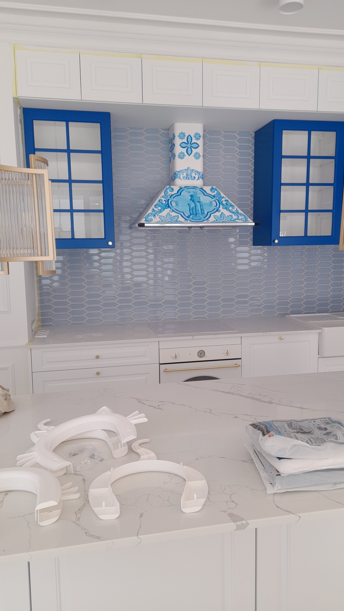 Nowoczesna kuchnia z niebieskimi akcentami: szafki z niebieskimi ramkami, dekoracyjny okap w stylu portugalskim, jasnoniebieskie płytki na ścianie, białe blaty z marmurowym wzorem. Na blacie...