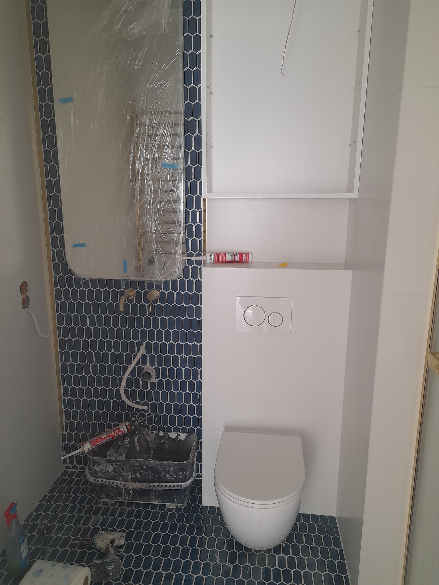 Łazienka w trakcie prac: ciemnoniebieska mozaika heksagonalna na ścianie i podłodze, wisząca toaleta, zabudowa z przyciskiem spłukiwania, grzejnik zakryty folią ochronną, widoczny pojemnik...