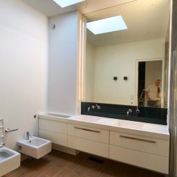 ROYAL BUD Kompleksowe Wykończenia wnętrz YURII TYMOSHCHUK - Nowoczesna łazienka z dużym lustrem z podświetleniem LED, białą szafką pod umywalką, bidetem i drewnopodobną podłogą. W lustrze odbija się osoba.