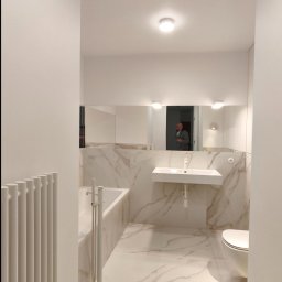 ROYAL BUD Kompleksowe Wykończenia wnętrz YURII TYMOSHCHUK - Jasna łazienka z marmurowymi płytkami, białą armaturą i grzejnikiem żeberkowym. W lustrze odbija się osoba robiąca zdjęcie.