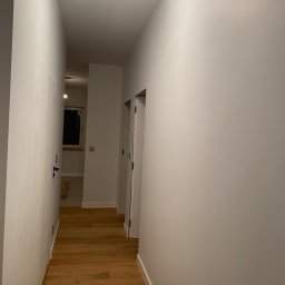 ROYAL BUD Kompleksowe Wykończenia wnętrz YURII TYMOSHCHUK - Pomalowany na biało korytarz z drewnianą podłogą, widoczna żarówka na suficie i drzwi po obu stronach, w głębi widoczne okno.