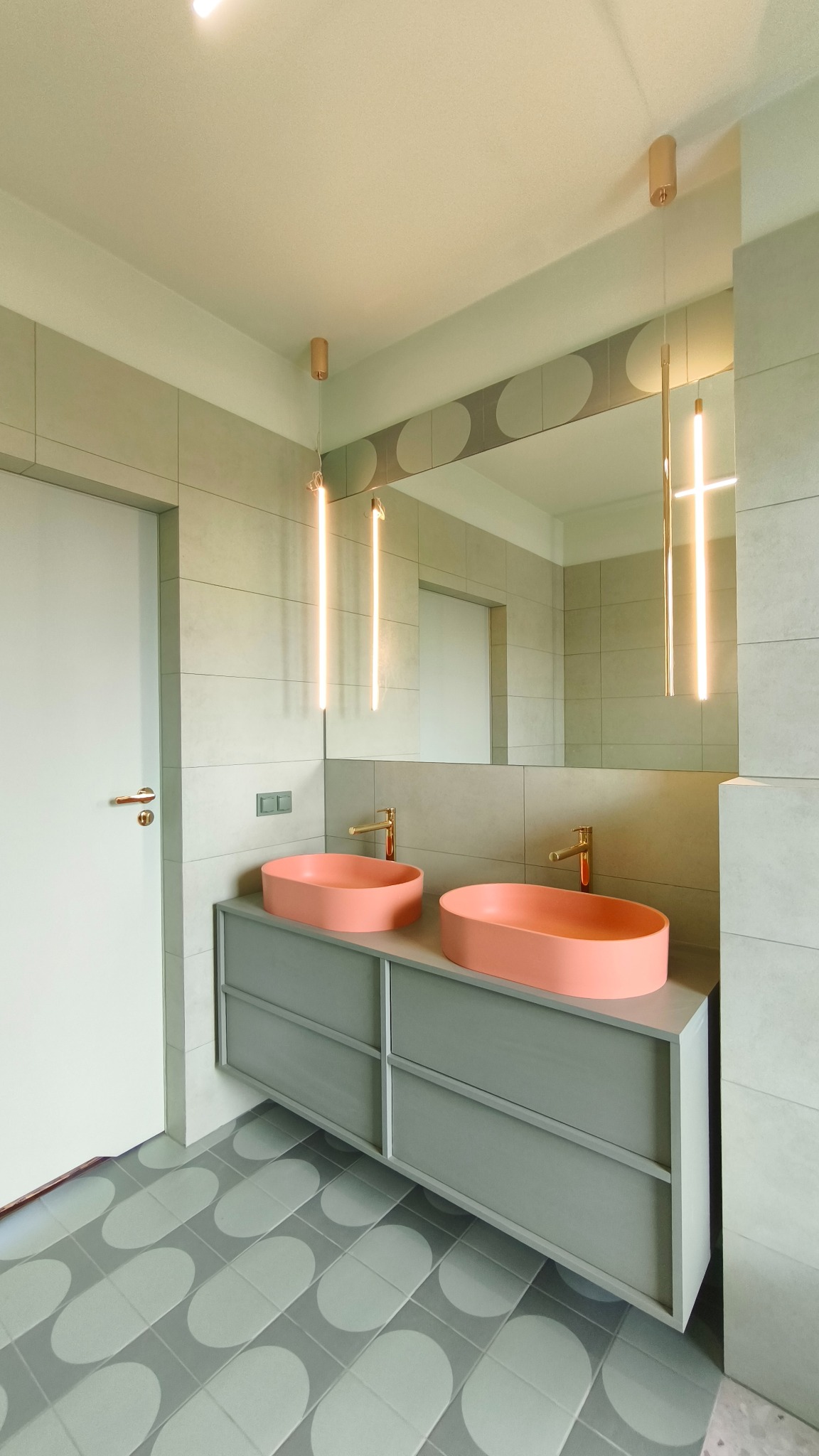 Nowoczesna łazienka z dwoma koralowymi umywalkami na szarej szafce, podświetlane lustro i wzorzysta podłoga. Minimalistyczny design z akcentem kolorystycznym.