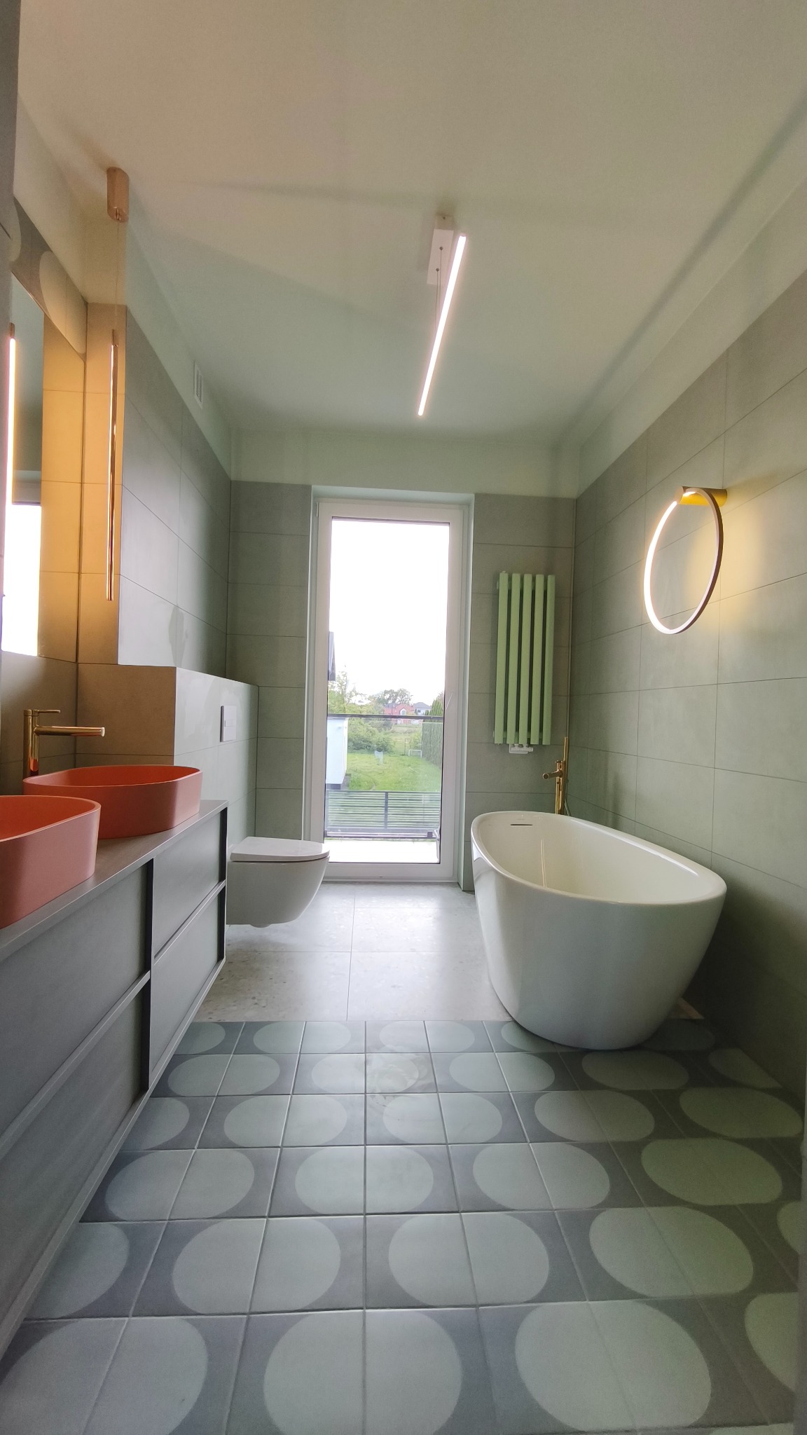 Nowoczesna łazienka w odcieniach szarości i zieleni z akcentami koralowymi: umywalki nablatowe, wolnostojąca wanna i okrągłe lustro z podświetleniem.