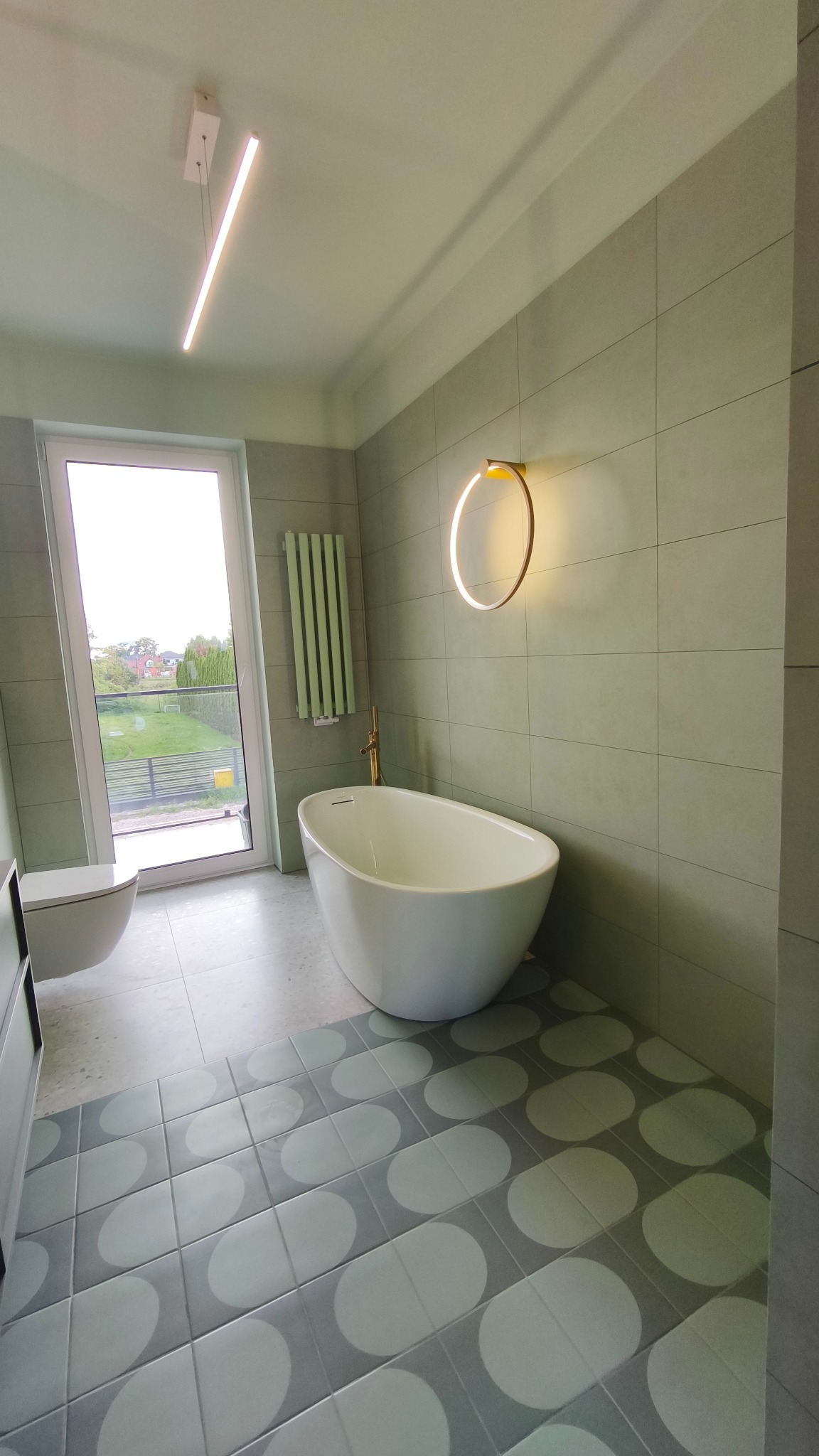 Nowoczesna łazienka w odcieniach szarości i zieleni z wolnostojącą wanną, okrągłym oświetleniem ściennym i grzejnikiem drabinkowym. Widok na zewnątrz przez okno.