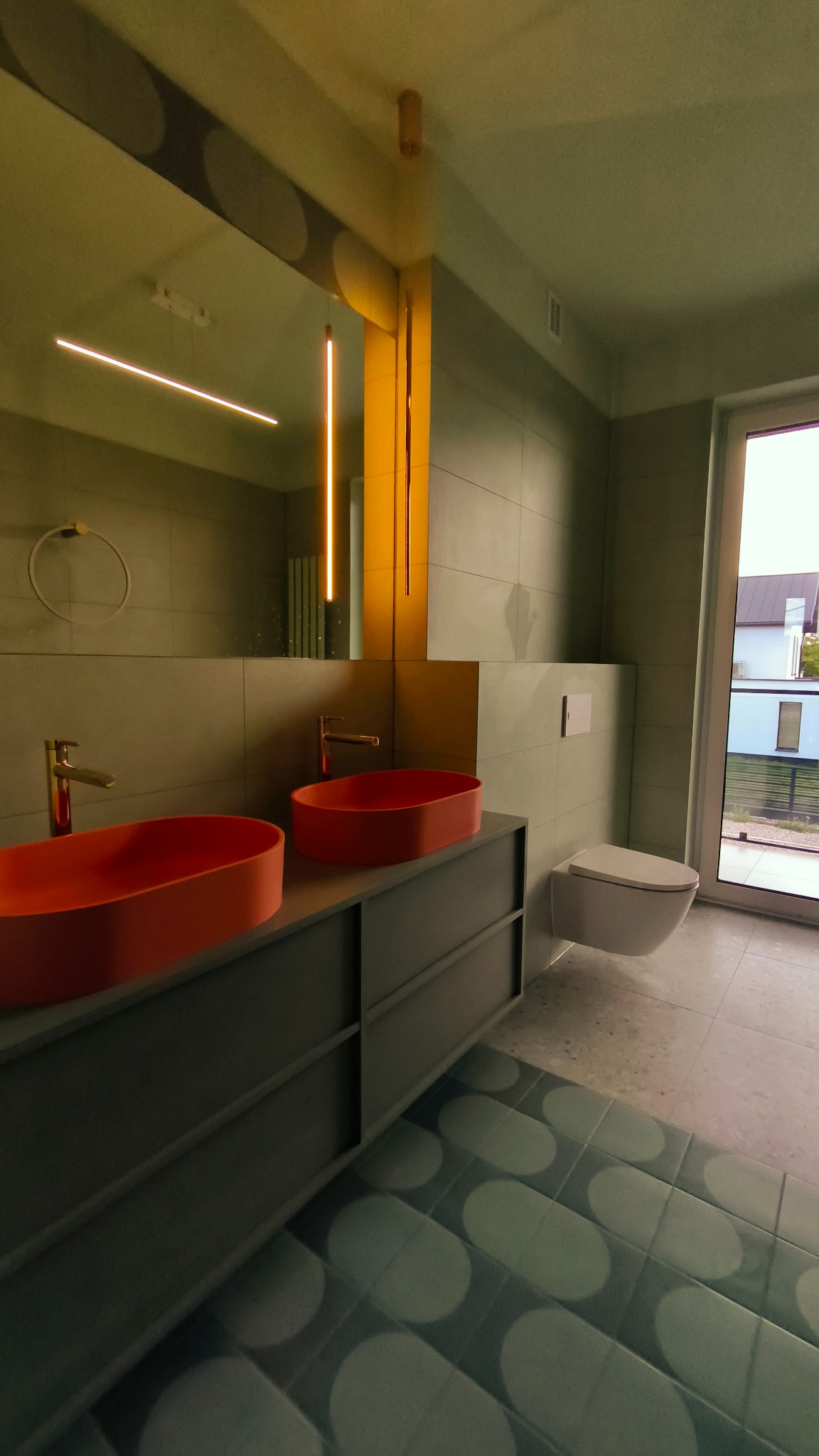 Nowoczesna łazienka z dwoma pomarańczowymi umywalkami na szarej szafce, podświetlane lustro i oryginalne płytki podłogowe w geometryczny wzór.