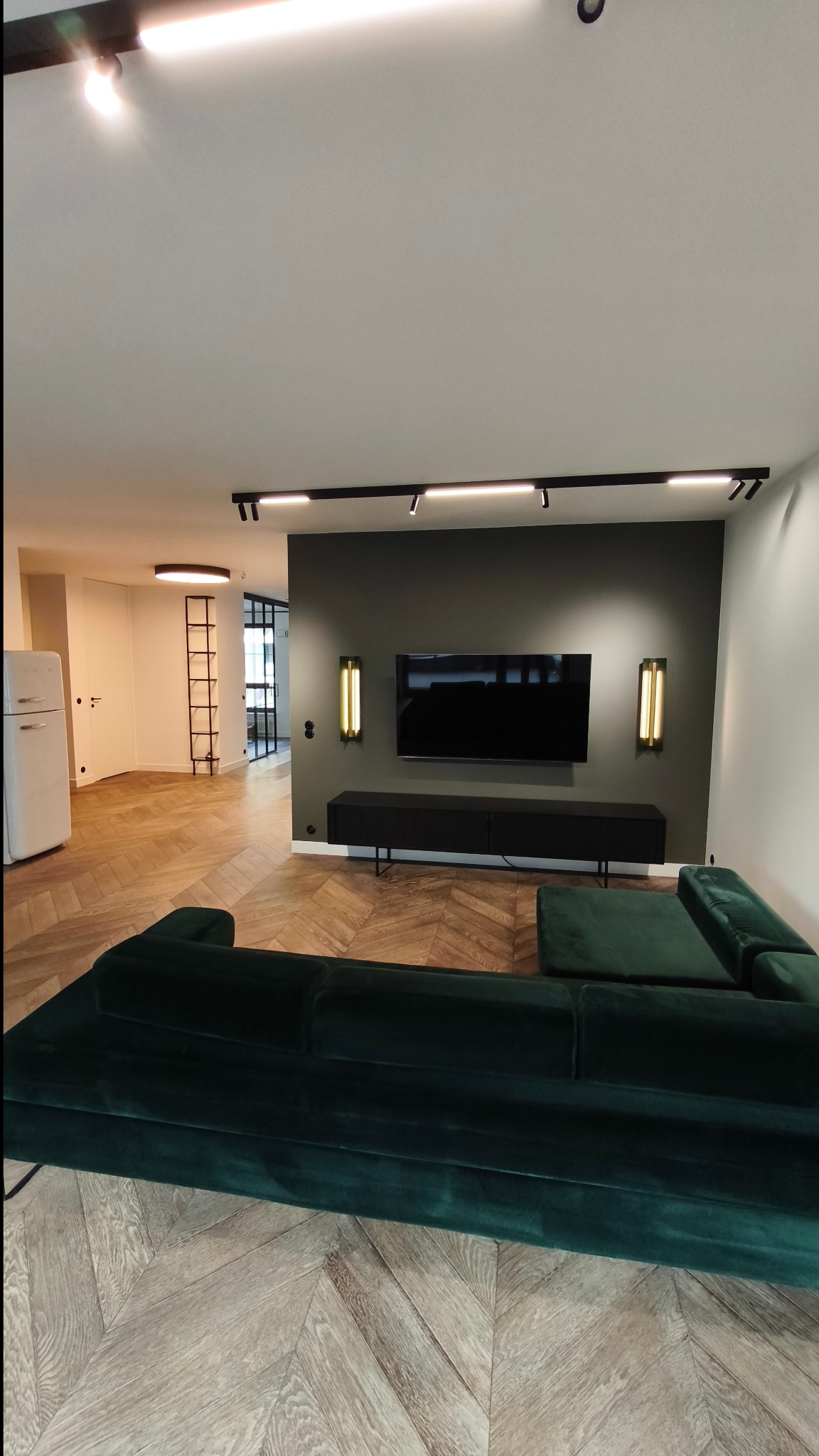 Salon po remoncie: parkiet w jodełkę, ciemnozielona sofa, czarna szafka RTV i telewizor na ścianie w kolorze grafitowym, oświetlenie szynowe.