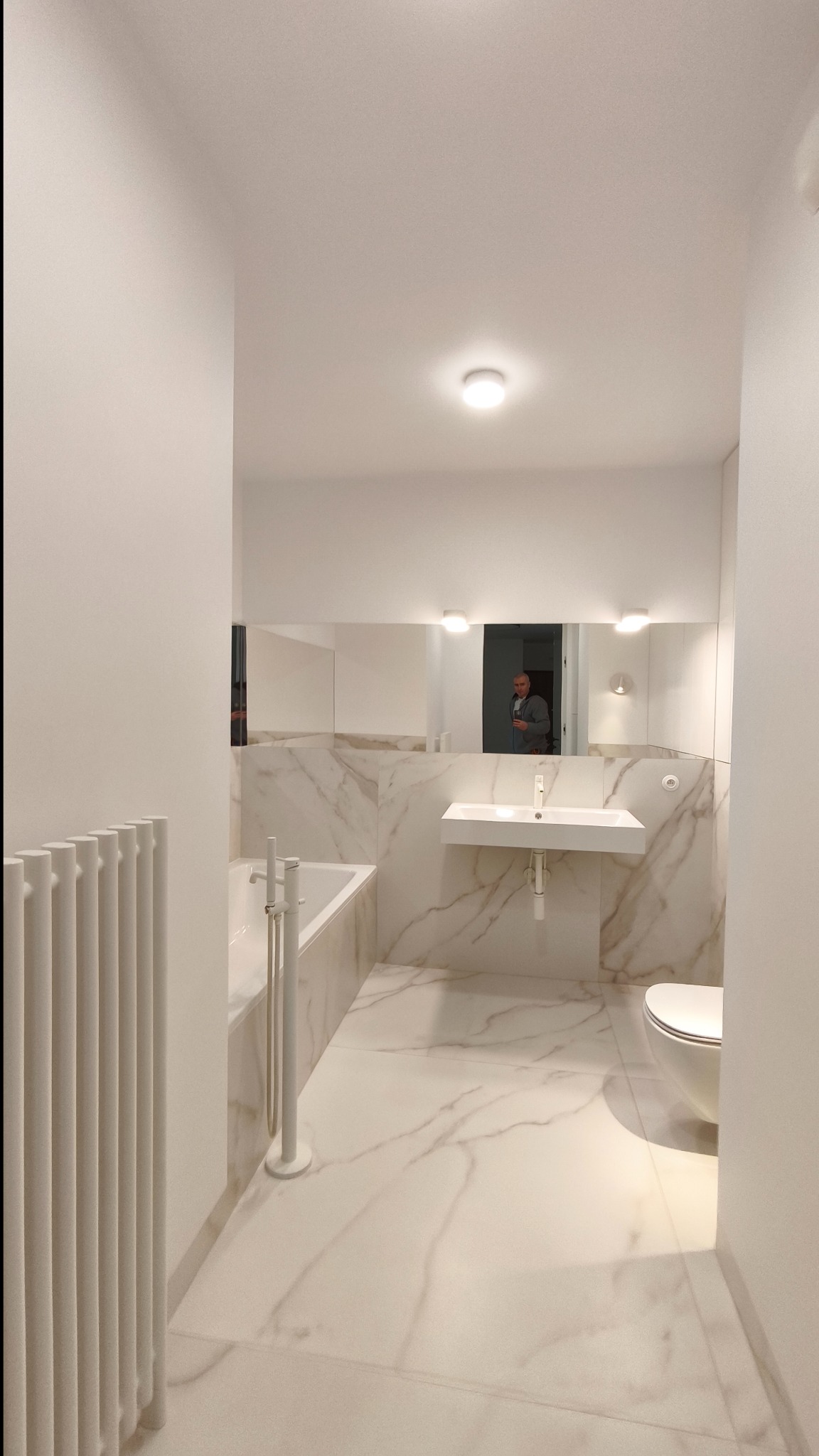 Jasna łazienka z marmurowymi płytkami, białą armaturą i grzejnikiem żeberkowym. W lustrze odbija się osoba robiąca zdjęcie.