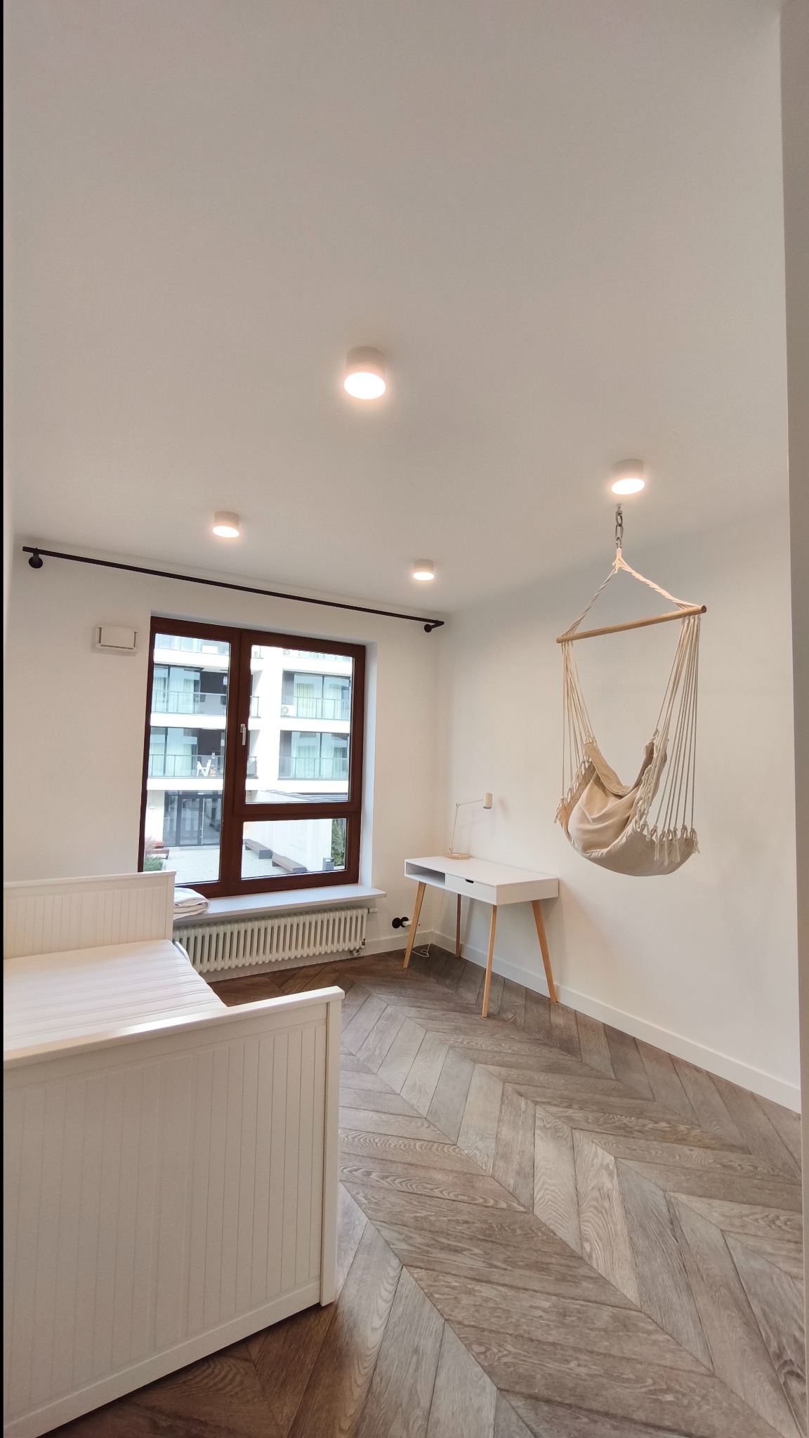 Jasny pokój z parkietem w jodełkę: białe ściany, okno z brązową ramą, białe biurko i łóżko, wiszący fotel hamakowy. Minimalistyczny design.