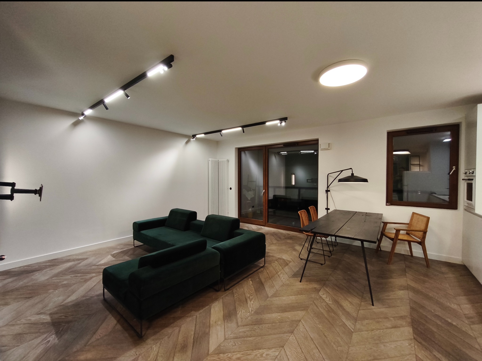 Salon z parkietem w jodełkę, zieloną sofą i nowoczesnym oświetleniem szynowym. Minimalistyczny design, jasne ściany, drewniane okno i drzwi przesuwne.