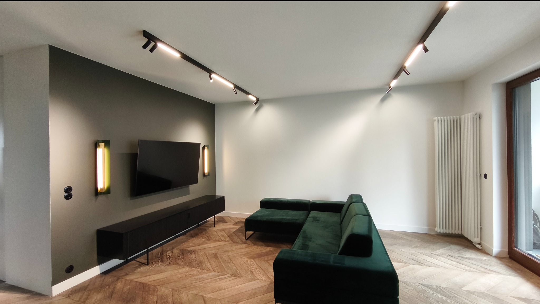 Salon po metamorfozie: parkiet w jodełkę, ciemnozielona sofa, ściana z TV i podłużne lampy ścienne. Nowoczesne oświetlenie punktowe na suficie.
