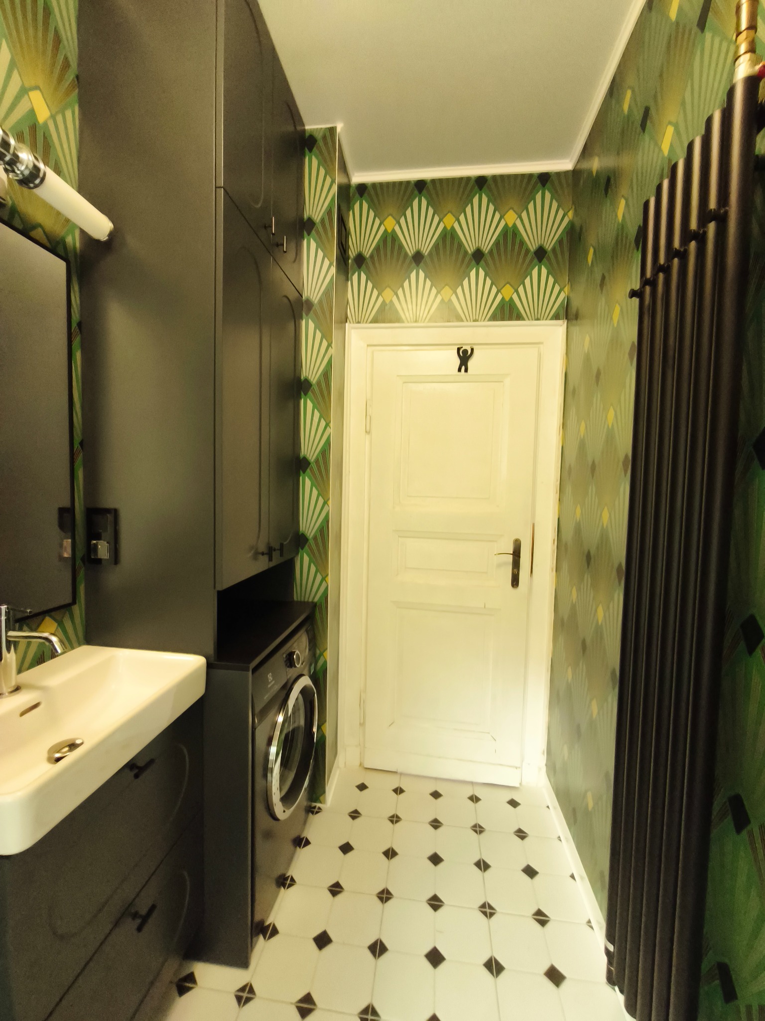 Nowoczesna łazienka z umywalką nablatową, szafką w kolorze grafitu, pralką i wzorzystą tapetą w stylu art déco. Czarno-biała podłoga w karo.