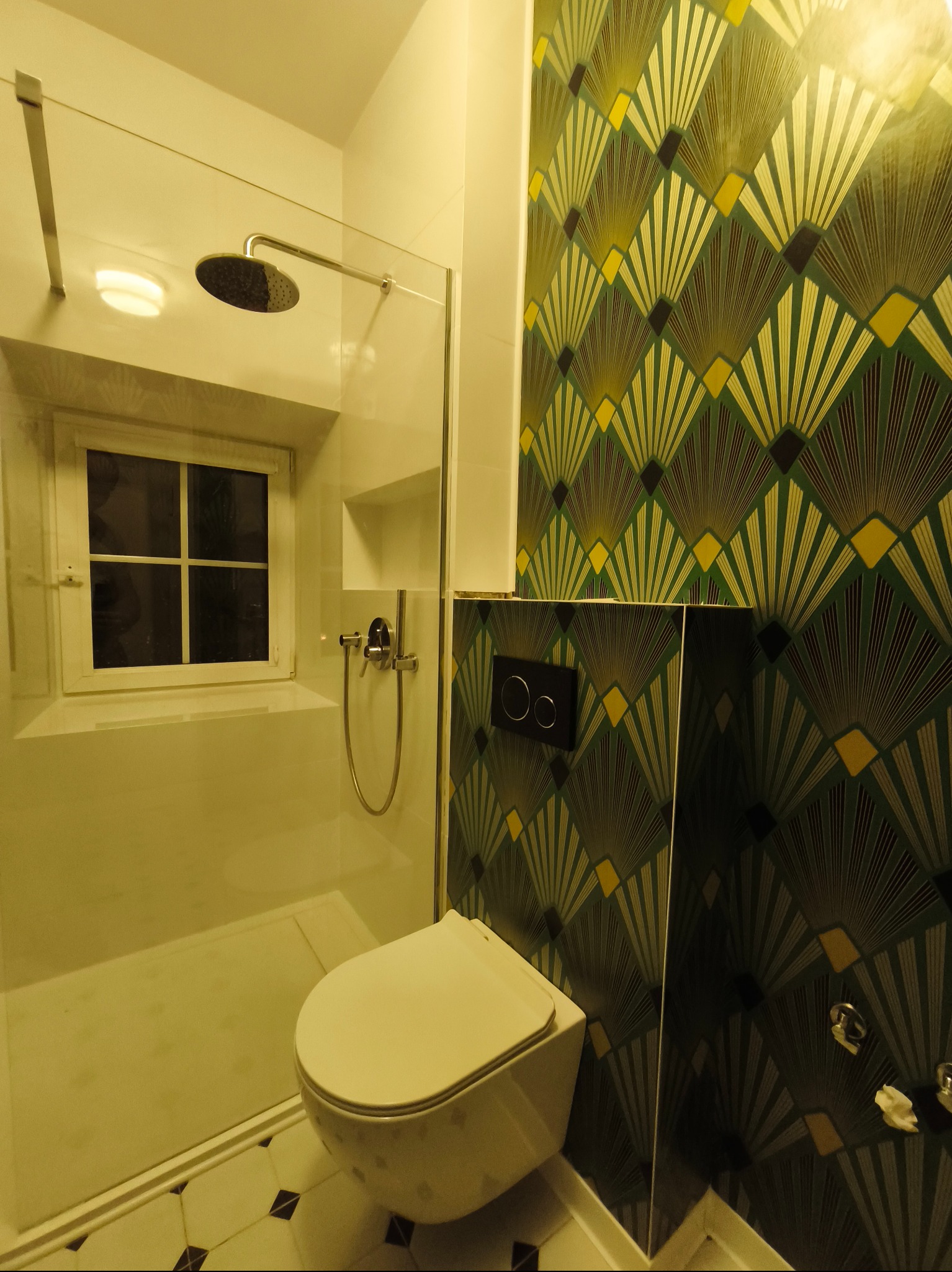 Wykończona łazienka w Warszawie z kabiną prysznicową i toaletą podwieszaną. Ściany zdobią tapety w geometryczne wzory w odcieniach zieleni i złota, nadając wnętrzu unikalny charakter.