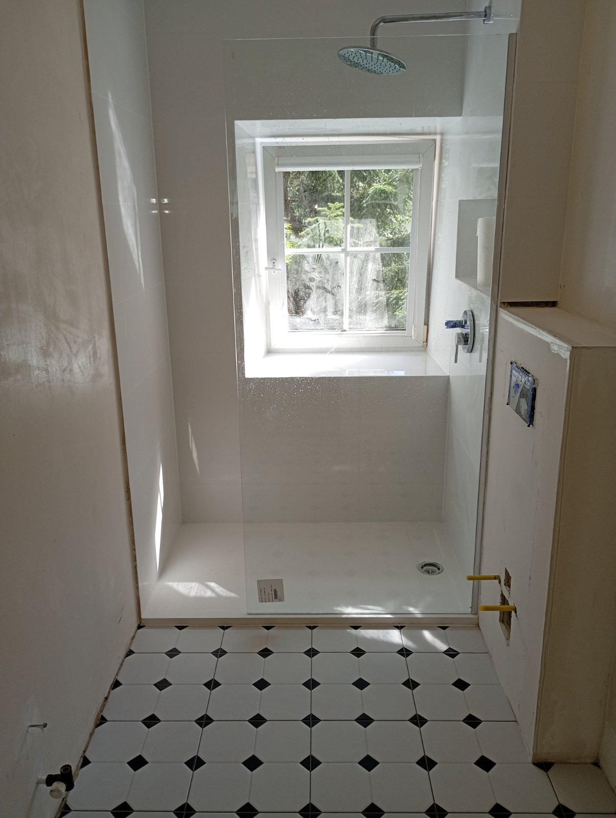 Biała kabina prysznicowa z oknem w nowoczesnej łazience. Widoczne biało-czarne płytki podłogowe i fragment ściany w trakcie wykończenia. Minimalistyczny design.