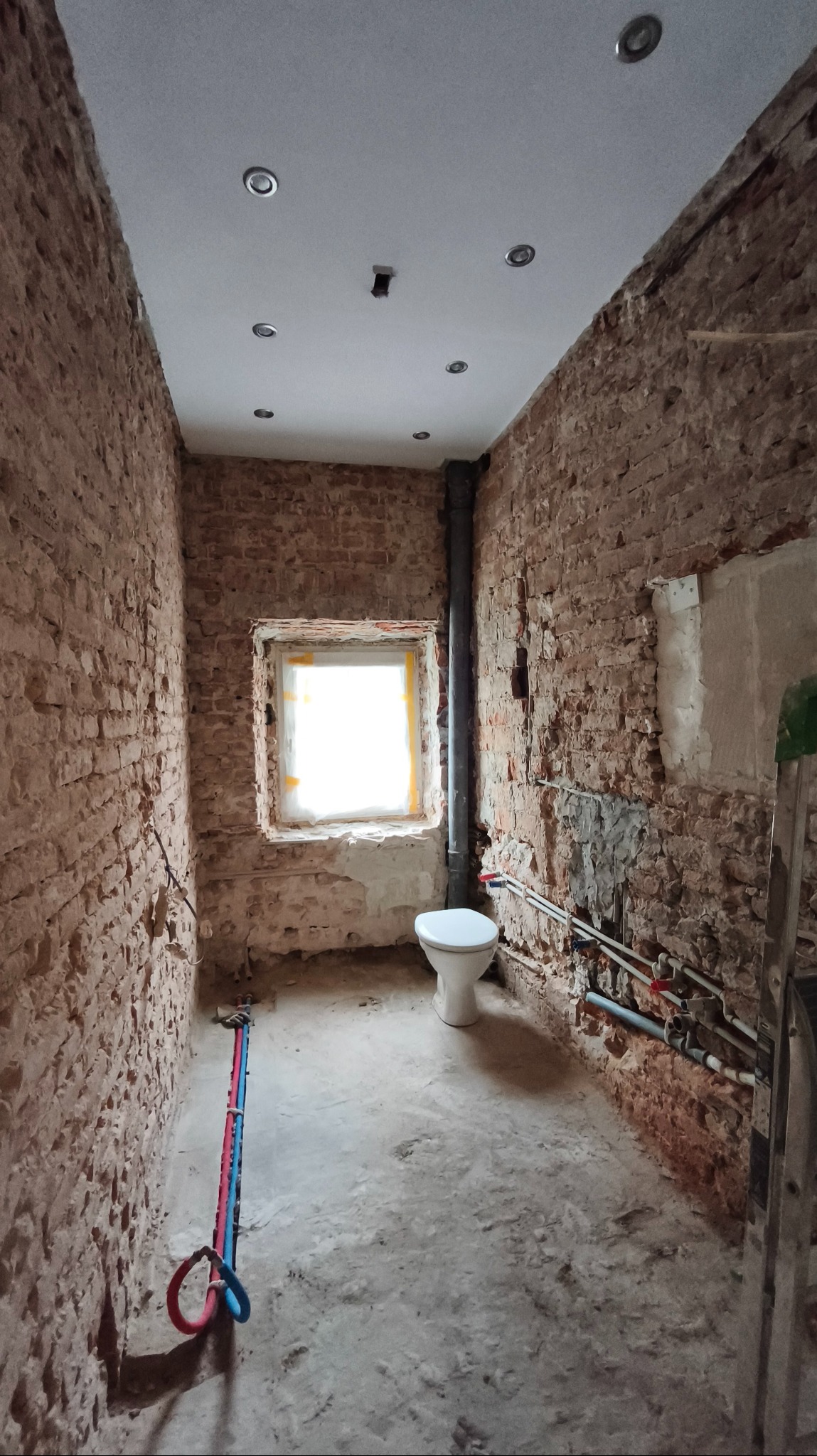 Łazienka w trakcie remontu: surowe ceglane ściany, częściowo wykończony sufit z oświetleniem, nowe rury i toaleta. Widoczne ślady prac remontowych.