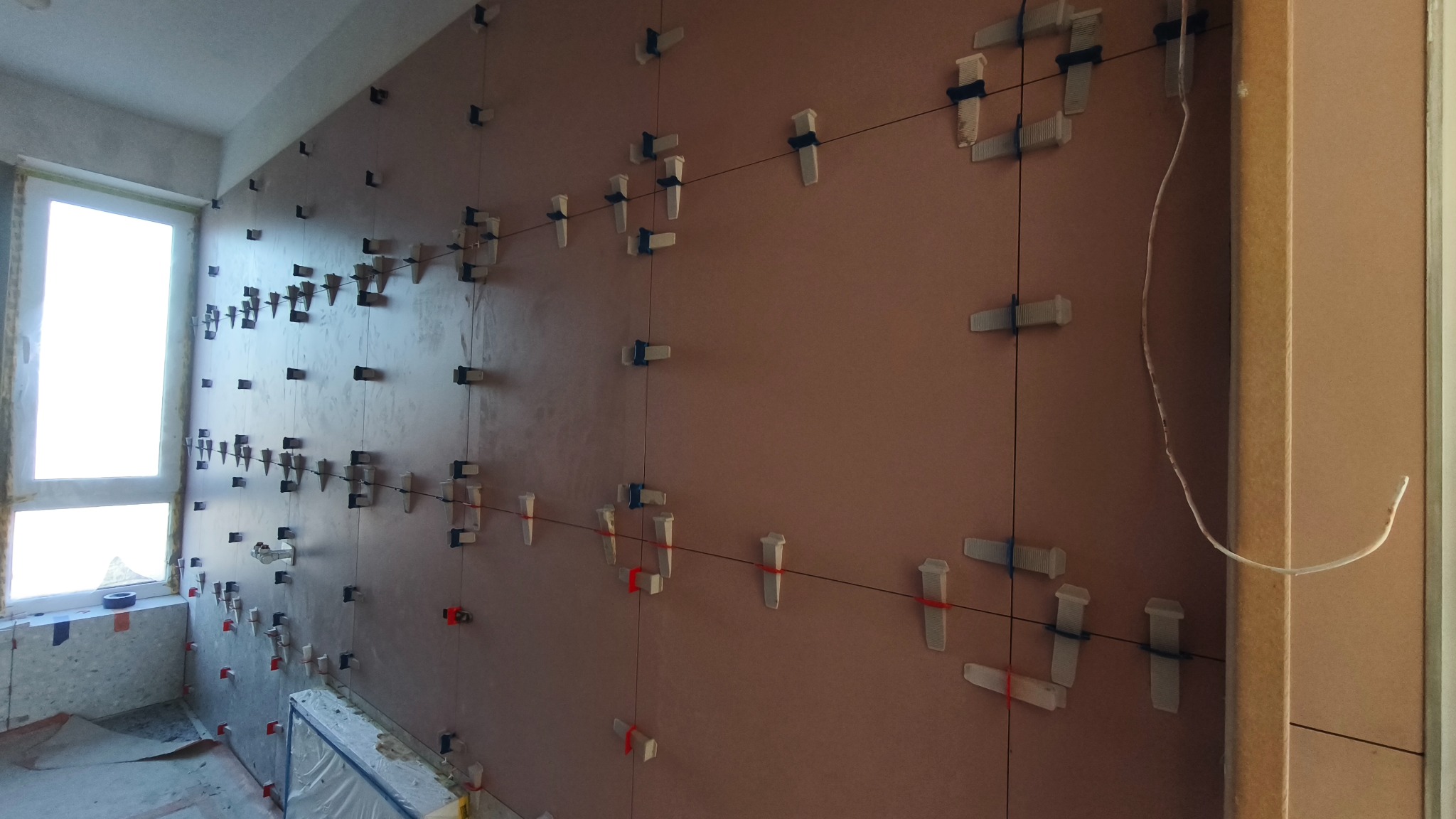 Ściana z zamocowanymi klipsami i sznurkami poziomującymi, przygotowana do układania płytek. Widoczne okno i fragment podłogi z zabezpieczeniami.