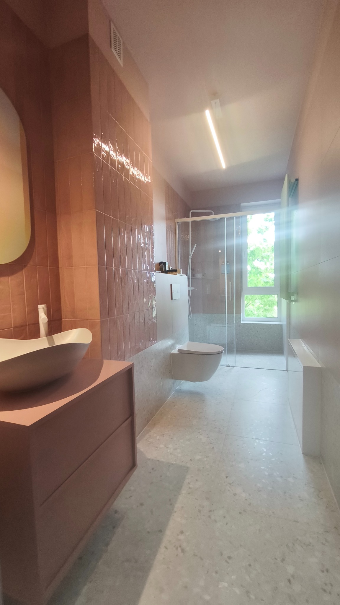 Nowoczesna łazienka w odcieniach różu i szarości z kabiną prysznicową, umywalką nablatową i podwieszaną toaletą. Minimalistyczny design, jasne wnętrze z oknem.