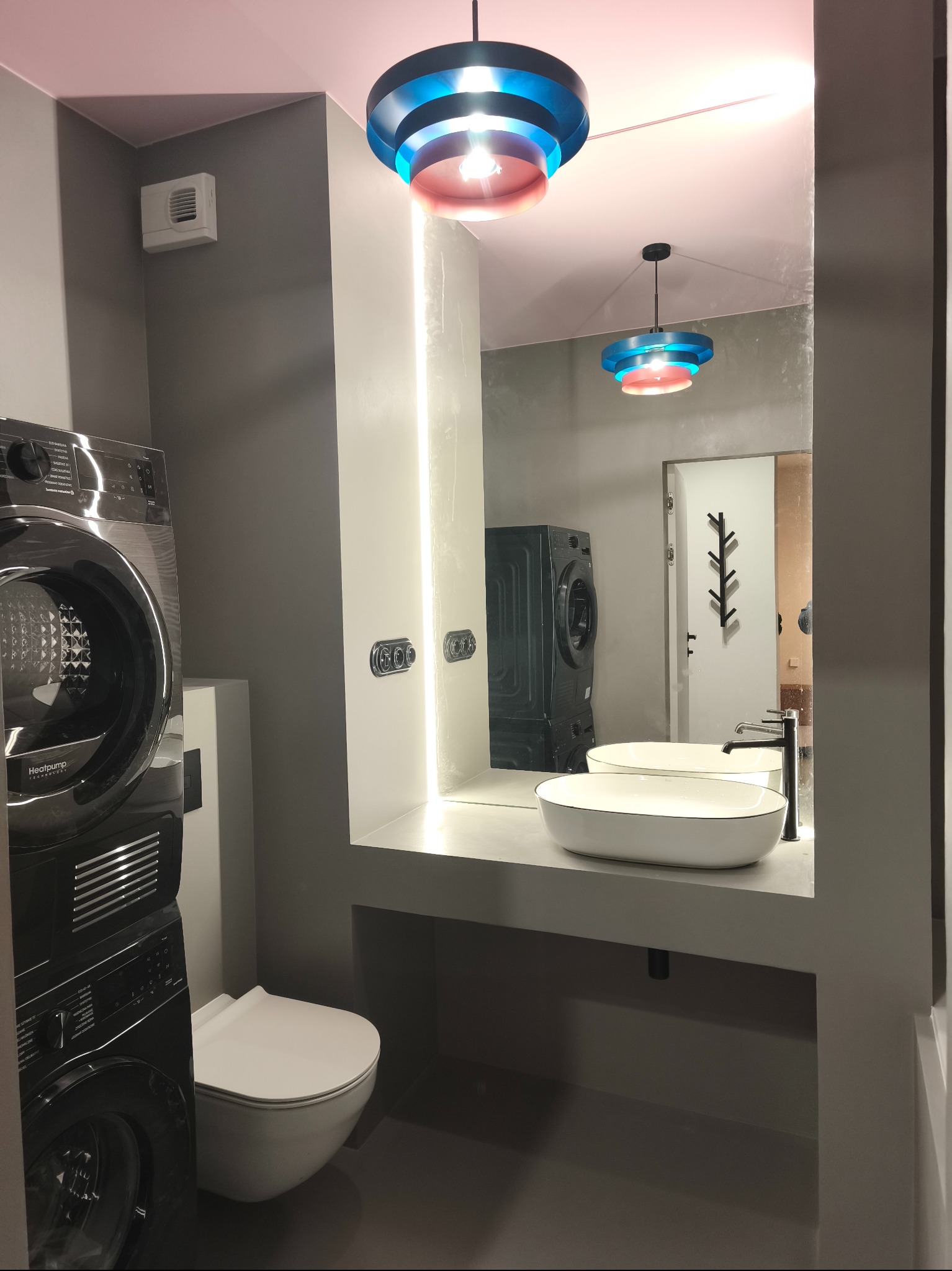 Nowoczesna łazienka w szarościach z pralką i suszarką w zabudowie, umywalką nablatową i podświetlanym lustrem. Kolorowa lampa dodaje akcentu.