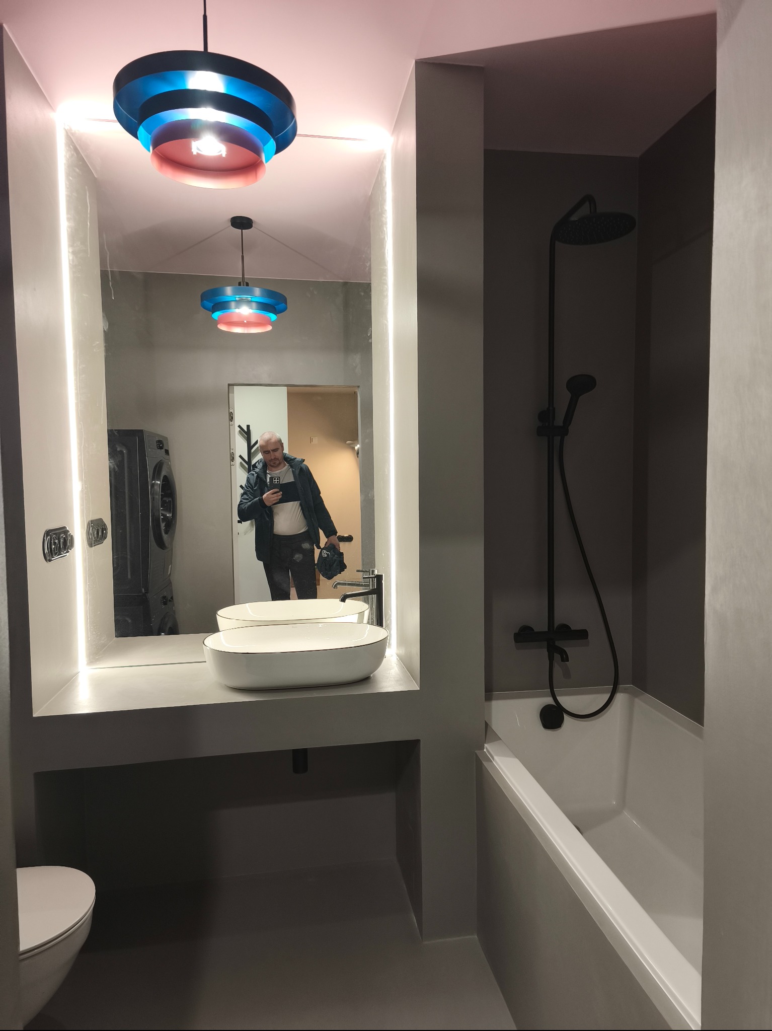 Nowoczesna łazienka w szarościach z podświetlanym lustrem i designerską lampą. W odbiciu widać osobę robiącą zdjęcie i pralkę. Minimalistyczny styl.