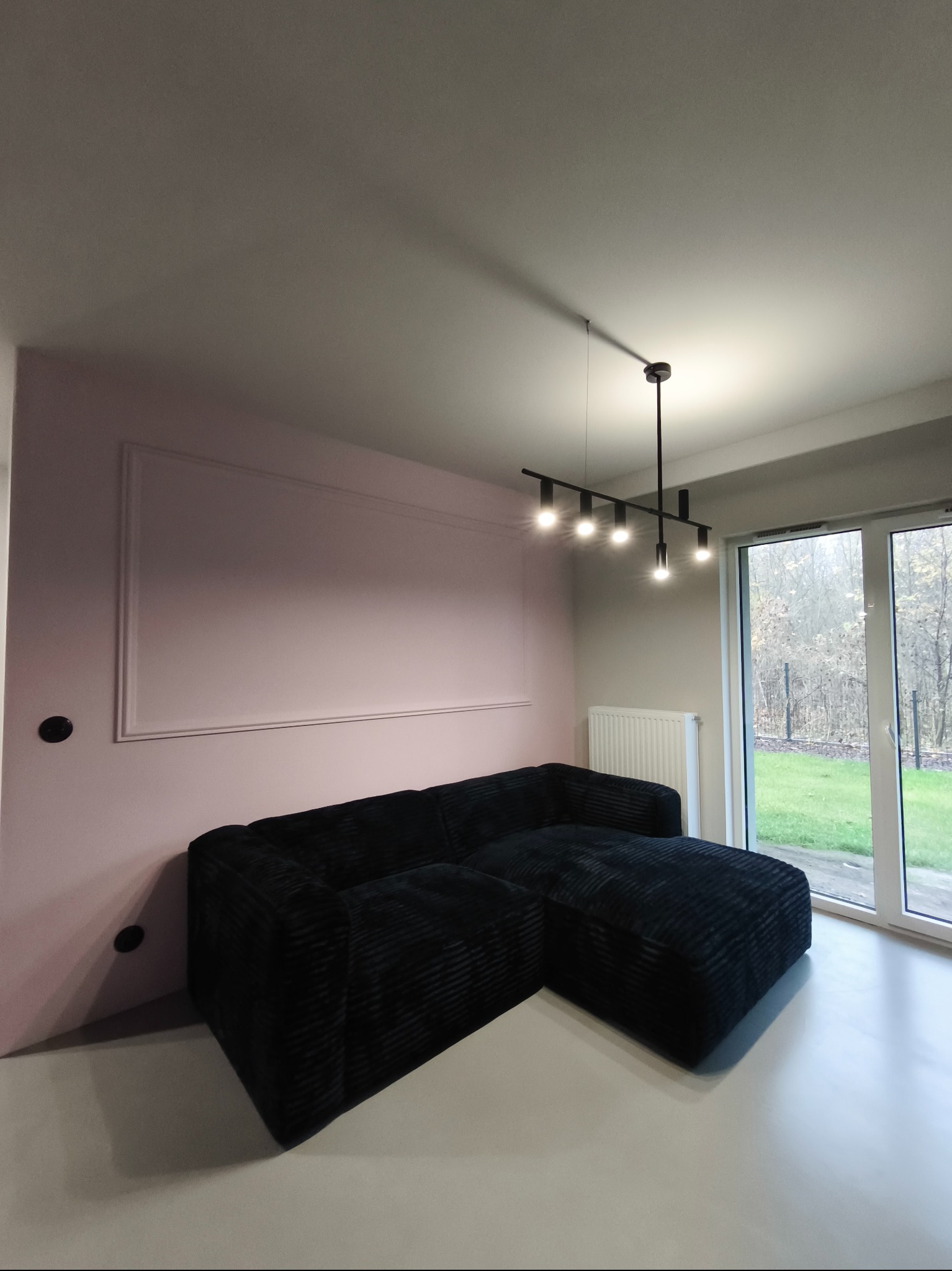Nowoczesny salon z czarną sofą narożną, różową ścianą z dekoracyjną ramą i lampą sufitową. Widok na ogród przez duże okno.