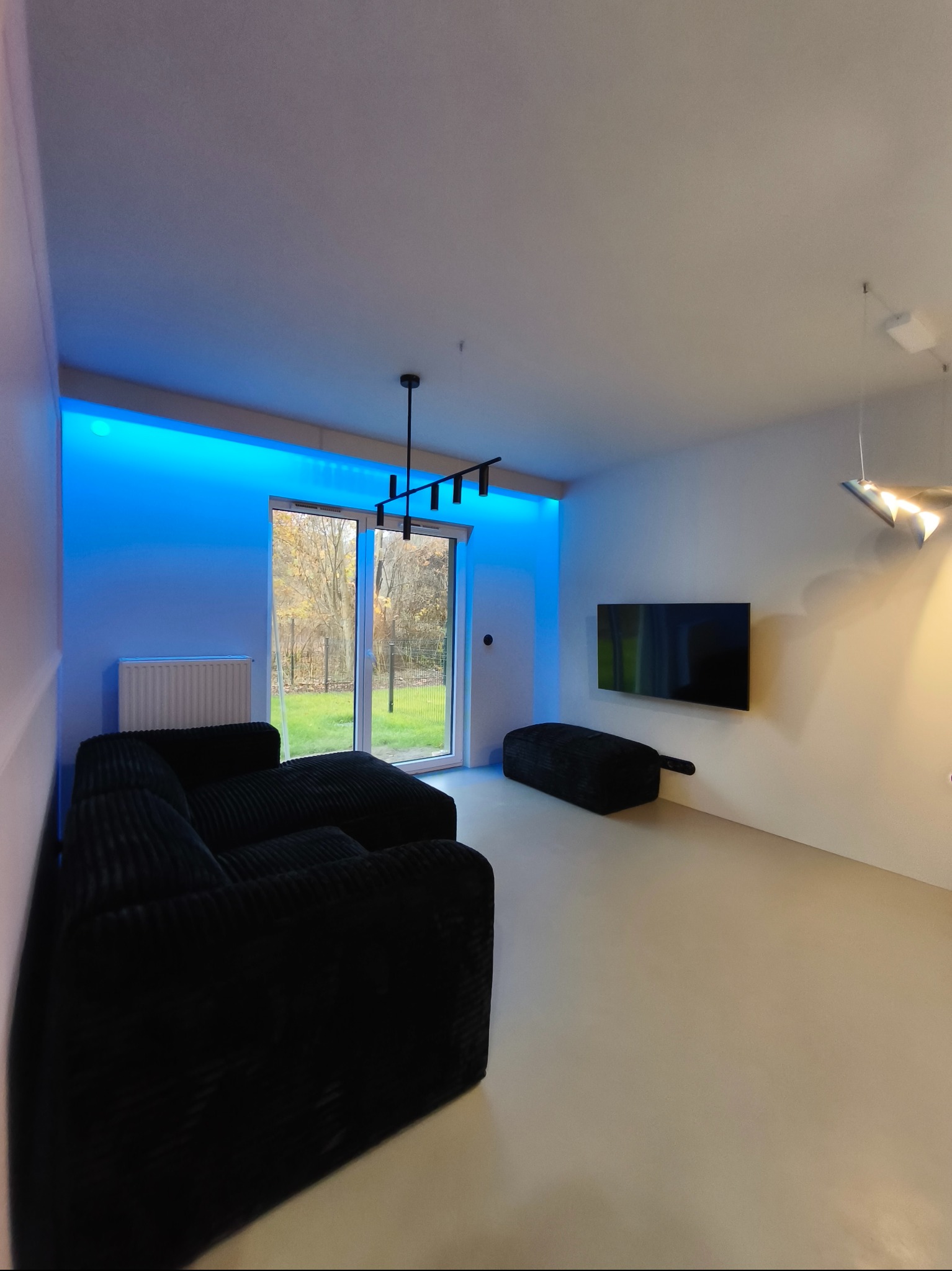 Minimalistyczny salon z czarną sofą i podświetleniem LED. Widok na ogród przez duże okno. Nowoczesne lampy i telewizor na ścianie. Jasna podłoga.