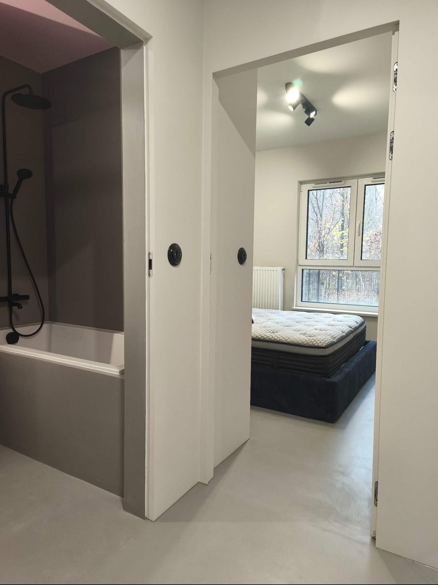 Nowoczesne wnętrze: widok na łazienkę z czarną armaturą i sypialnię z dużym oknem, przez otwarte białe drzwi. Minimalistyczny design, szara kolorystyka, jednolite podłogi.