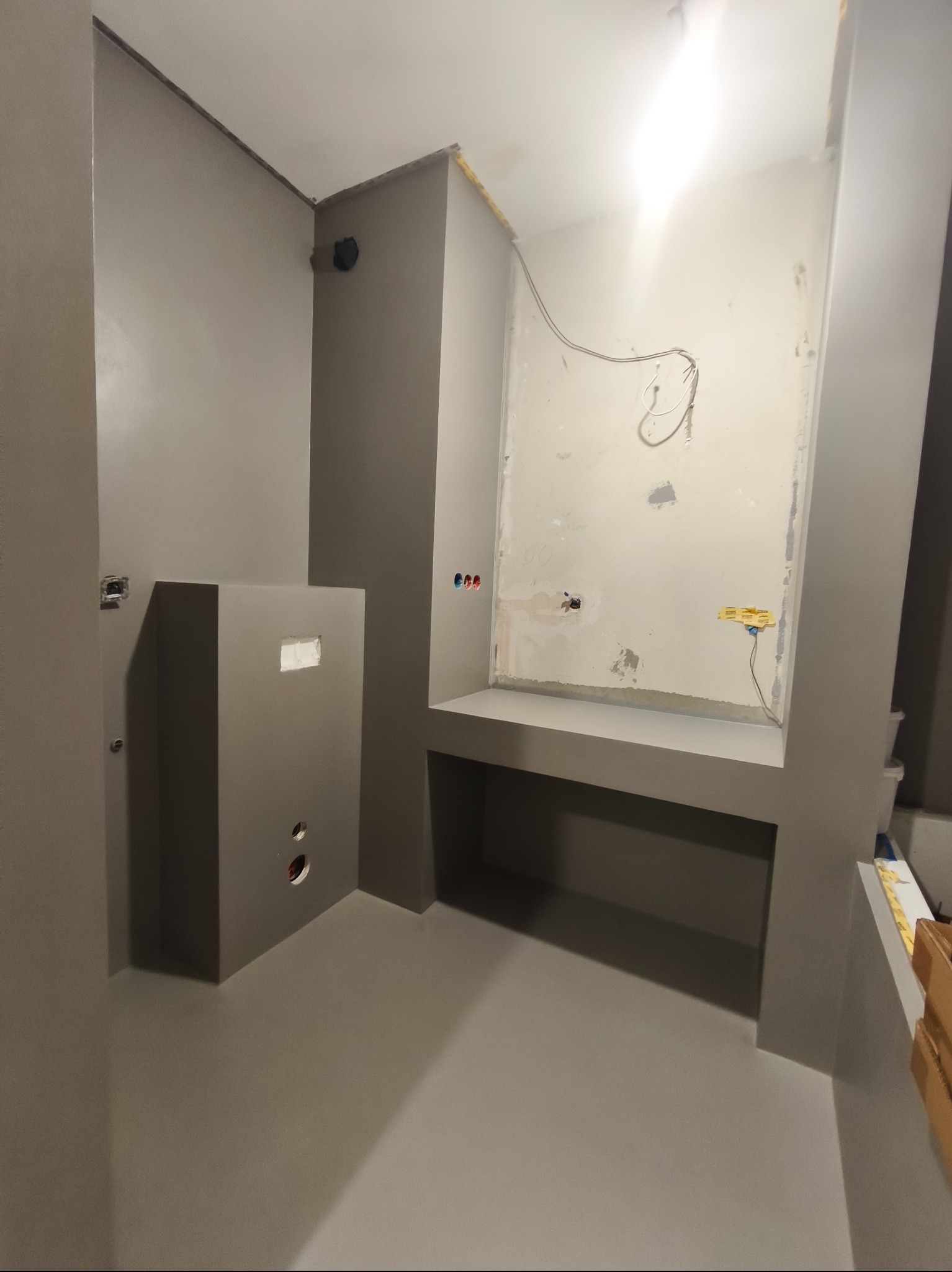 Szara łazienka w trakcie wykończenia: ściany z gładzią, przygotowane instalacje, surowy stan przed montażem armatury. Widoczne kable i otwory na gniazdka.