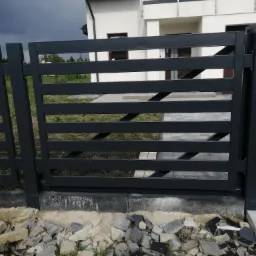 Nowoczesne, poziome ogrodzenie z metalowych profili w kolorze grafitowym, z widoczną furtką i fragmentem domu w tle, teren wokół ogrodzenia częściowo wyłożony kostką, a częściowo gruzem.