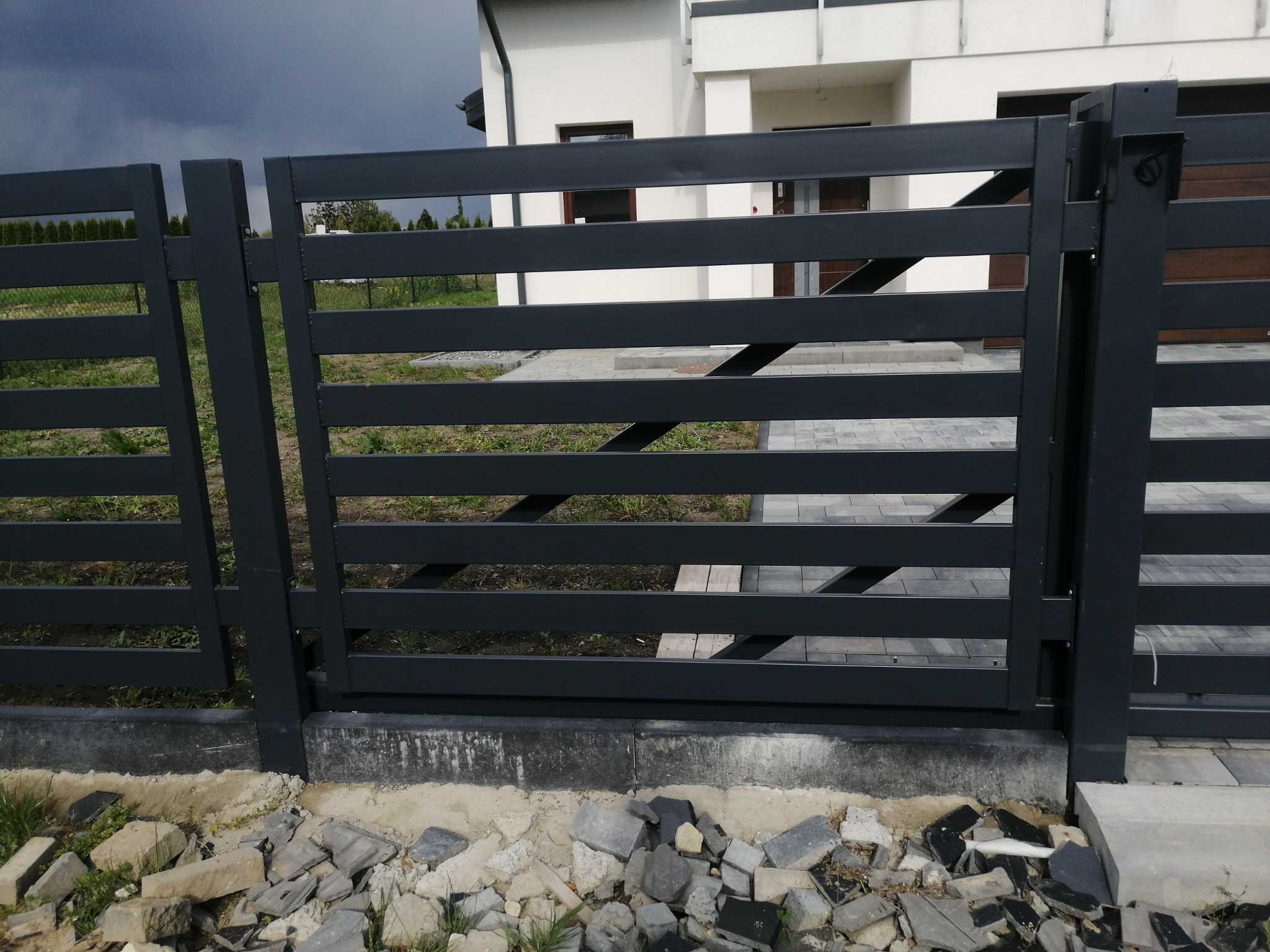Nowoczesne, poziome ogrodzenie z metalowych profili w kolorze grafitowym, z widoczną furtką i fragmentem domu w tle, teren wokół ogrodzenia częściowo wyłożony kostką, a częściowo gruzem.