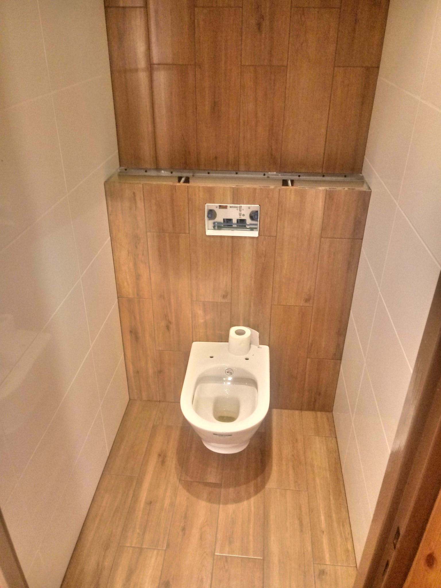 Wykończona toaleta w małej łazience z drewnopodobnymi płytkami na podłodze i ścianie za toaletą, jasne płytki na pozostałych ścianach, widoczny fragment drewnianej futryny drzwi.